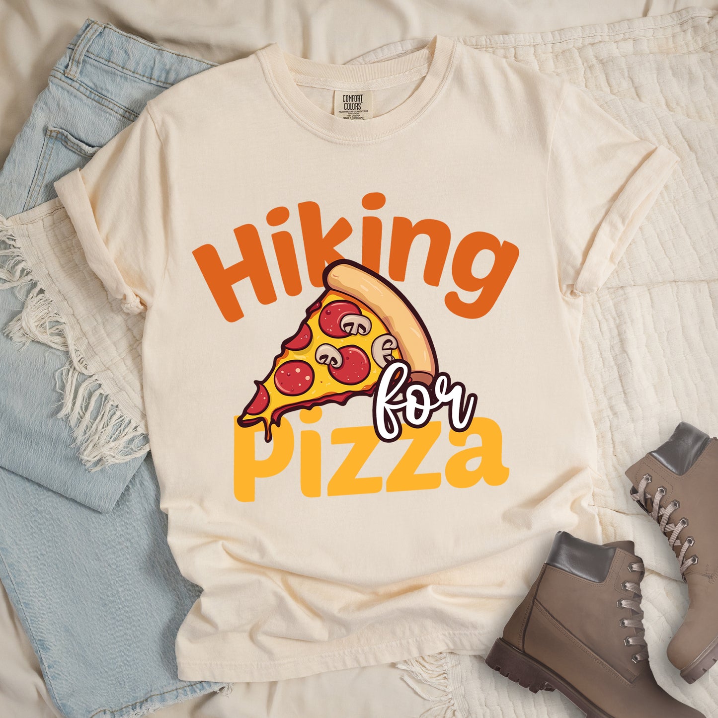 Ivory (Cream White) t-shirt with yellow and orange text reading "Hiking for PIZZA". "Hiking" appears in orange curved text at the top, "for" in small white script in the middle, and "PIZZA" in bold yellow letters at the bottom. The centerpiece is a detailed illustration of a pizza slice with pepperoni and mushroom toppings on a yellow cheese background with tan crust. Features a classic crew neck and casual rolled sleeves, styled alongside light wash denim and brown hiking boots.