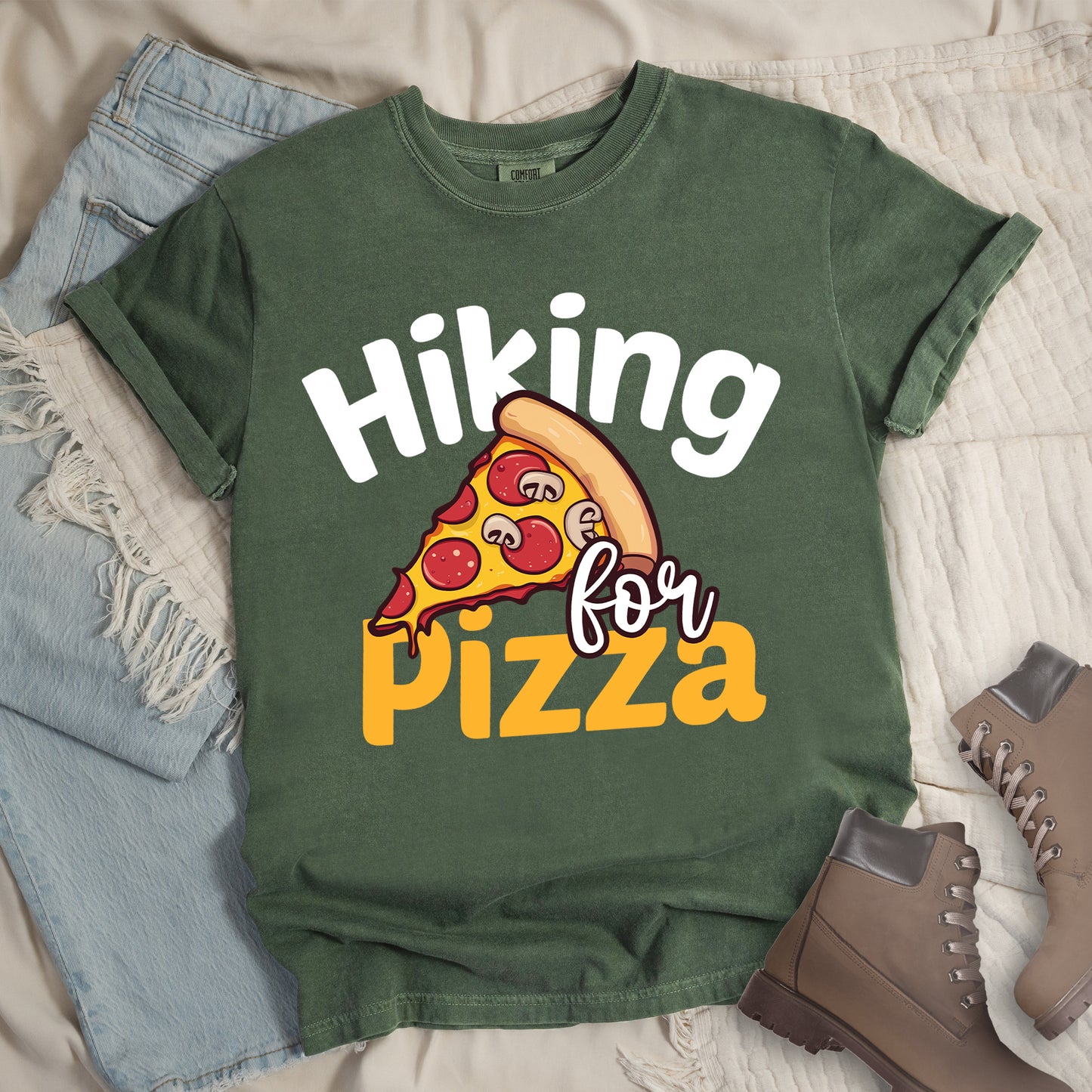 Moss (Dark Green) t-shirt with white and orange text reading "Hiking for PIZZA". "Hiking" appears in white curved text at the top, "for" in small white script in the middle, and "PIZZA" in bold orange letters at the bottom. The centerpiece is a detailed illustration of a pizza slice with pepperoni and mushroom toppings on a yellow cheese background with tan crust. Features a classic crew neck and casual rolled sleeves, styled alongside light wash denim and brown hiking boots.