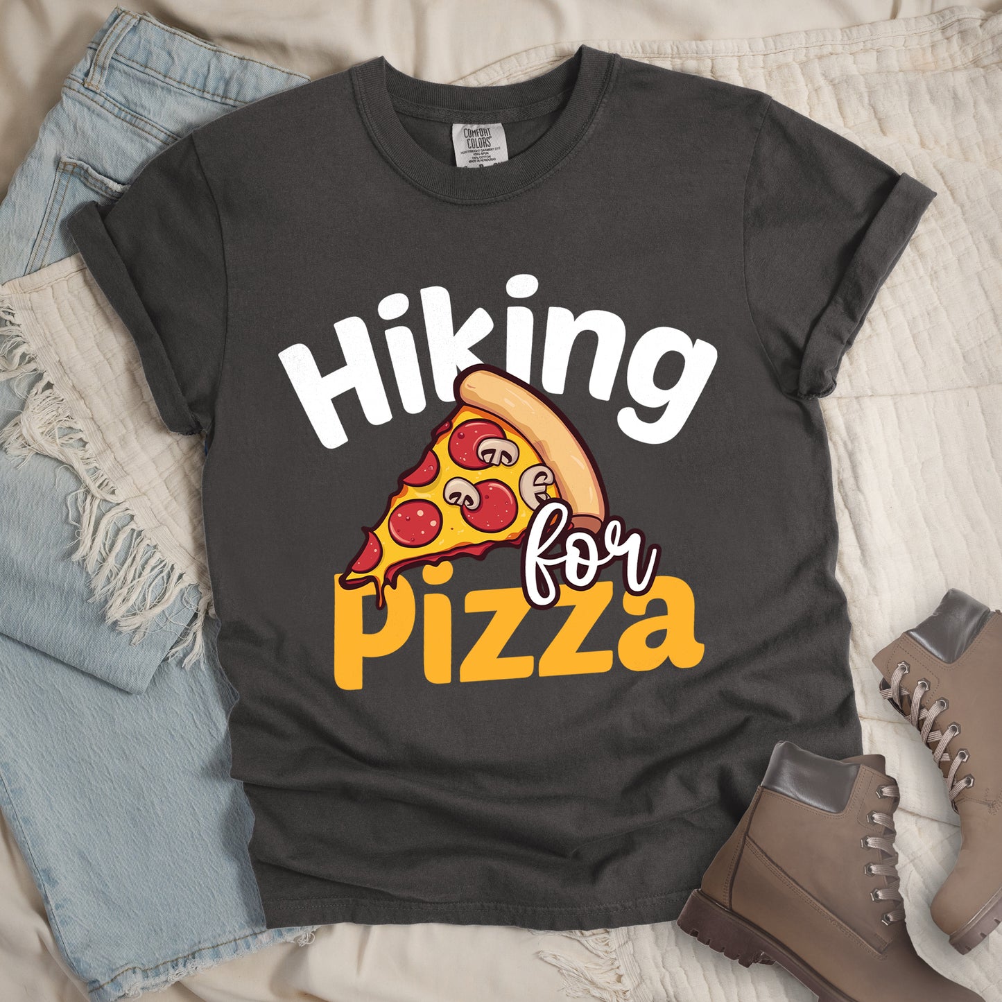Pepper (Dark Gray) t-shirt with white and orange text reading "Hiking for PIZZA". "Hiking" appears in white curved text at the top, "for" in small white script in the middle, and "PIZZA" in bold orange letters at the bottom. The centerpiece is a detailed illustration of a pizza slice with pepperoni and mushroom toppings on a yellow cheese background with tan crust. Features a classic crew neck and casual rolled sleeves, styled alongside light wash denim and brown hiking boots.