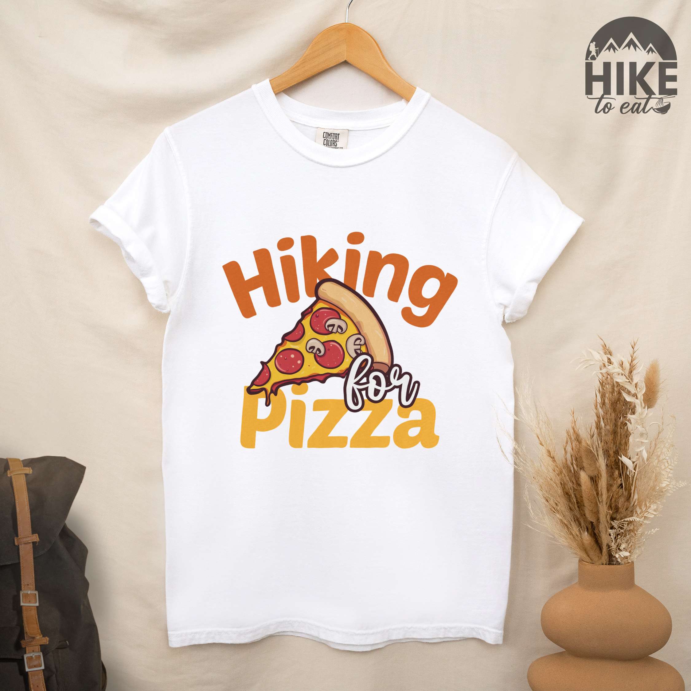 Hanging "Hiking for pizza" colored vintage tee with bold typography, illustrated pizza slice, and backpacker for pizza lover.