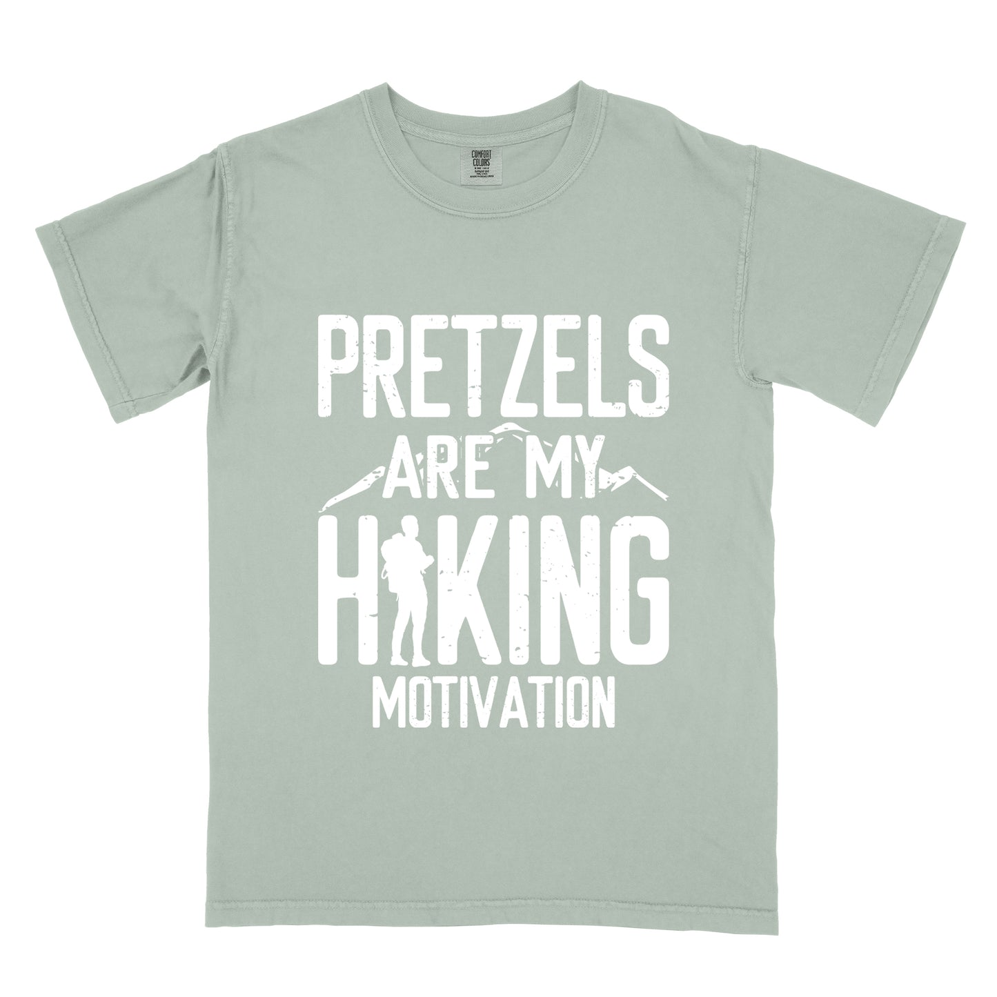 Bay (Light Gray) t-shirt with white distressed text reading "PRETZELS ARE MY HIKING MOTIVATION". "PRETZELS" appears at the top, "ARE MY" in the middle with small mountain silhouettes behind it, and "HIKING" in large letters with a hiker silhouette integrated into the letter "H". "MOTIVATION" appears at the bottom.