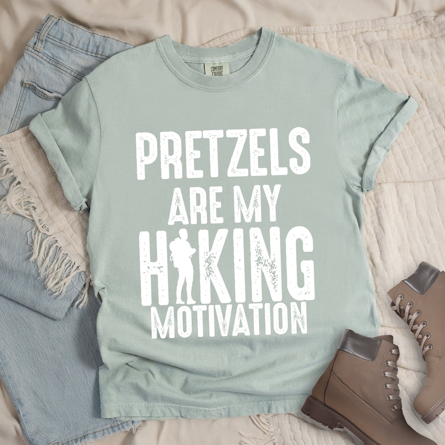 Bay (Light Gray) t-shirt with white distressed text reading "PRETZELS ARE MY HIKING MOTIVATION". "PRETZELS" appears at the top, "ARE MY" in the middle, and "HIKING" in large letters with a hiker silhouette integrated into the letter "I". "MOTIVATION" appears at the bottom. Features a classic crew neck and casual rolled sleeves, styled alongside light wash denim and brown hiking boots.