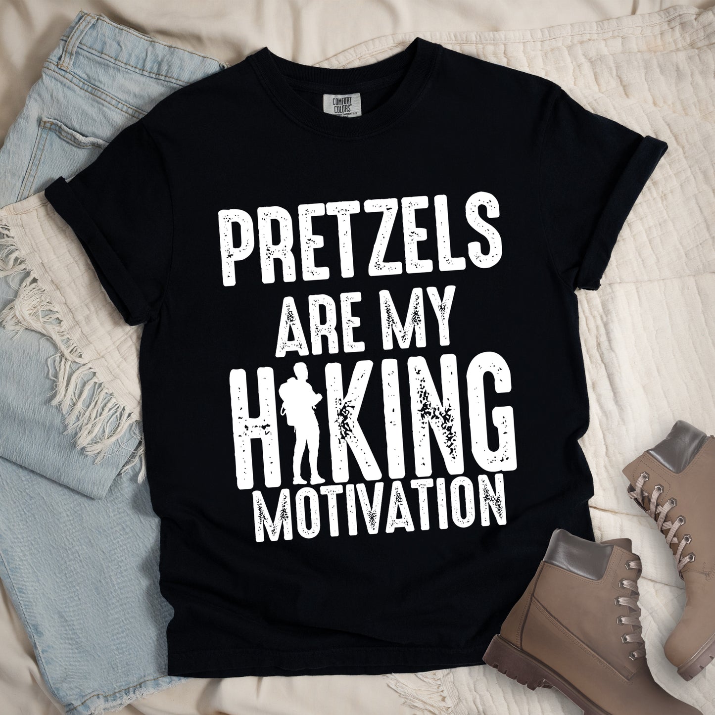 Black t-shirt with white distressed text reading "PRETZELS ARE MY HIKING MOTIVATION". "PRETZELS" appears at the top, "ARE MY" in the middle, and "HIKING" in large letters with a hiker silhouette integrated into the letter "I". "MOTIVATION" appears at the bottom. Features a classic crew neck and casual rolled sleeves, styled alongside light wash denim and brown hiking boots.