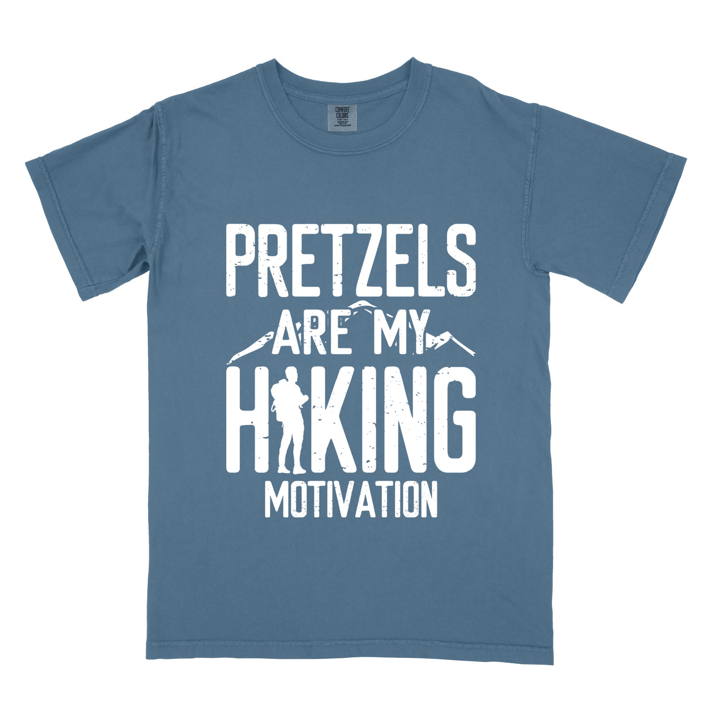 Blue Jean (Denim Blue) t-shirt with white distressed text reading "PRETZELS ARE MY HIKING MOTIVATION". "PRETZELS" appears at the top, "ARE MY" in the middle with small mountain silhouettes behind it, and "HIKING" in large letters with a hiker silhouette integrated into the letter "H". "MOTIVATION" appears at the bottom.