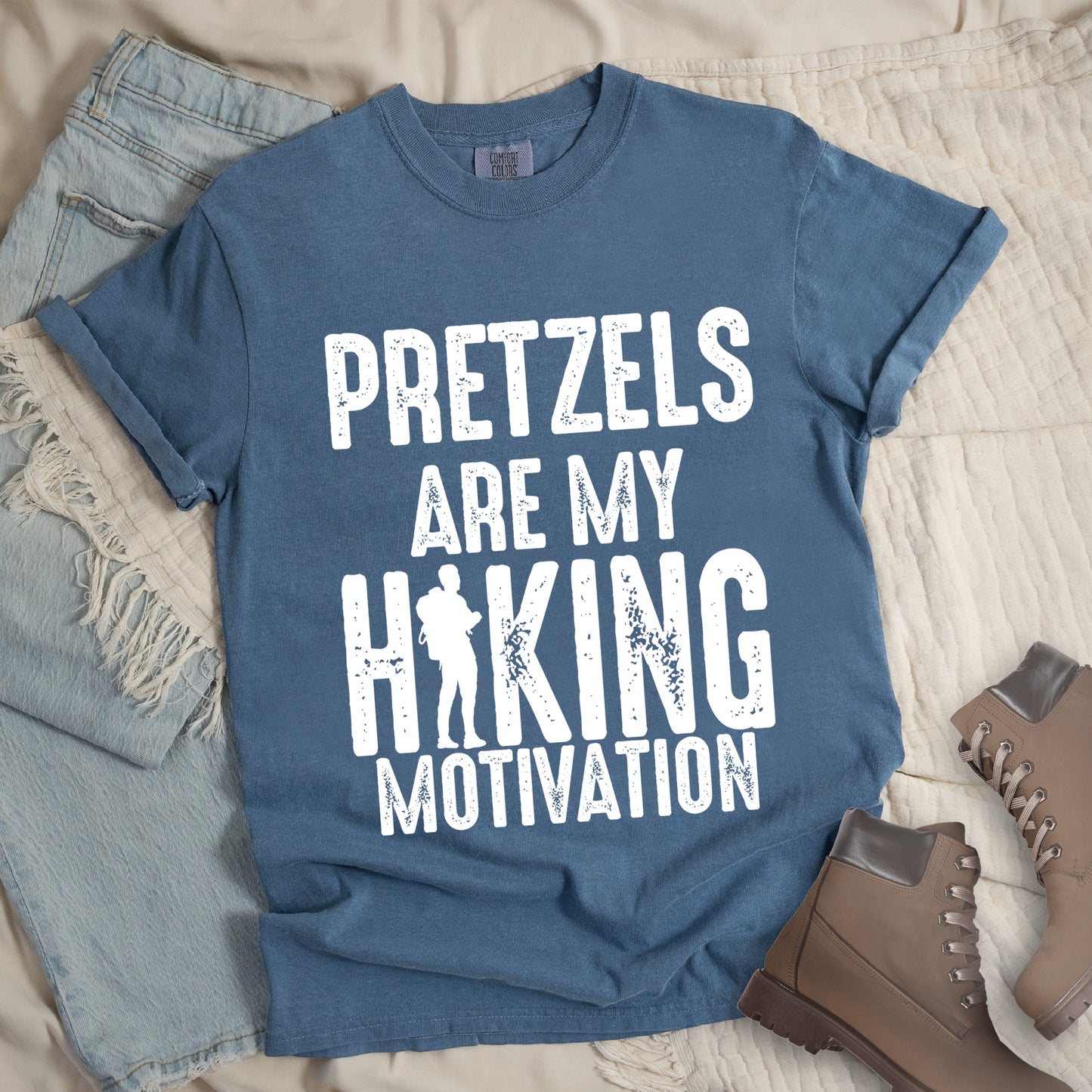 Blue Jean (Denim Blue) t-shirt with white distressed text reading "PRETZELS ARE MY HIKING MOTIVATION". "PRETZELS" appears at the top, "ARE MY" in the middle, and "HIKING" in large letters with a hiker silhouette integrated into the letter "I". "MOTIVATION" appears at the bottom. Features a classic crew neck and casual rolled sleeves, styled alongside light wash denim and brown hiking boots.