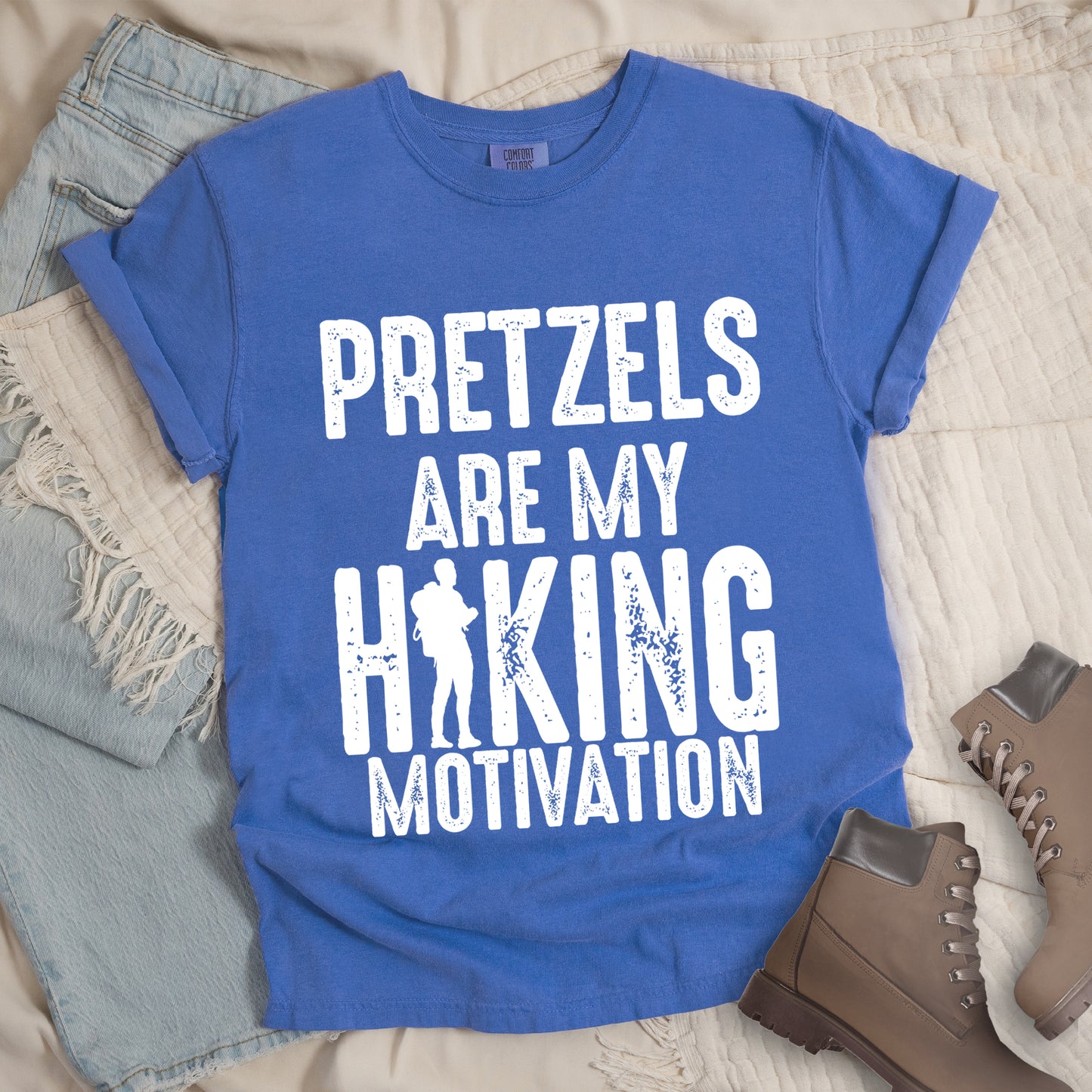 Flo Blue (Bright Blue) t-shirt with white distressed text reading "PRETZELS ARE MY HIKING MOTIVATION". "PRETZELS" appears at the top, "ARE MY" in the middle, and "HIKING" in large letters with a hiker silhouette integrated into the letter "I". "MOTIVATION" appears at the bottom. Features a classic crew neck and casual rolled sleeves, styled alongside light wash denim and brown hiking boots.