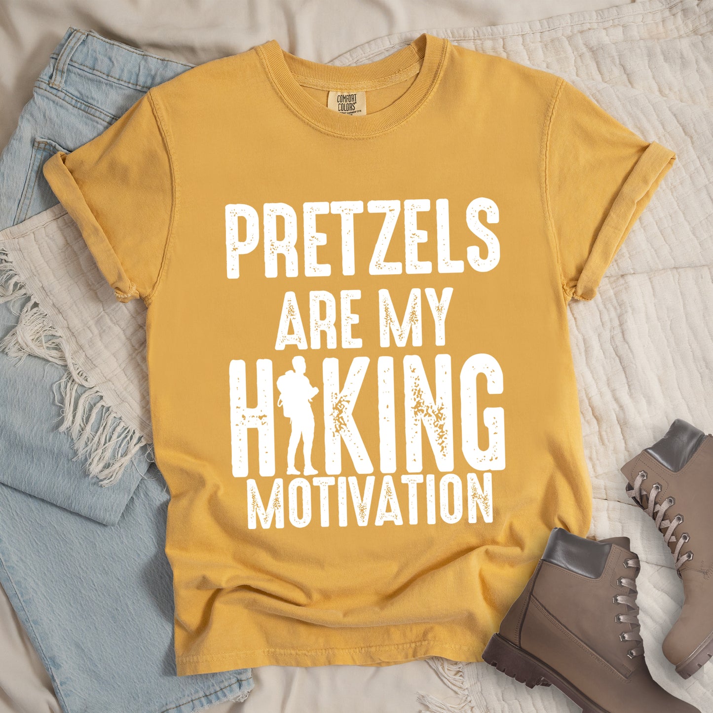 Mustard (Yellow) t-shirt with white distressed text reading "PRETZELS ARE MY HIKING MOTIVATION". "PRETZELS" appears at the top, "ARE MY" in the middle, and "HIKING" in large letters with a hiker silhouette integrated into the letter "I". "MOTIVATION" appears at the bottom. Features a classic crew neck and casual rolled sleeves, styled alongside light wash denim and brown hiking boots.
