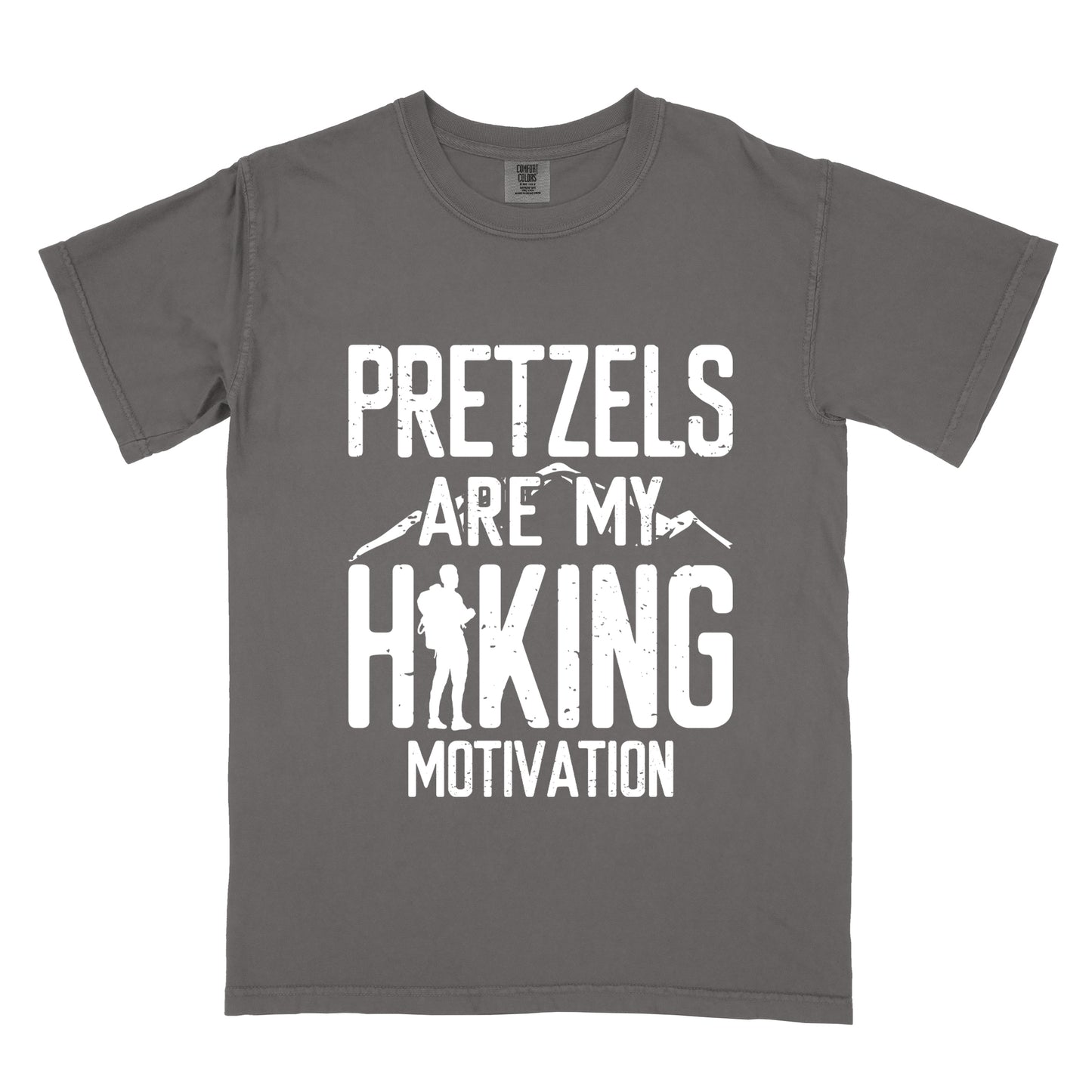 Pepper (Dark Gray) t-shirt with white distressed text reading "PRETZELS ARE MY HIKING MOTIVATION". "PRETZELS" appears at the top, "ARE MY" in the middle with small mountain silhouettes behind it, and "HIKING" in large letters with a hiker silhouette integrated into the letter "H". "MOTIVATION" appears at the bottom.