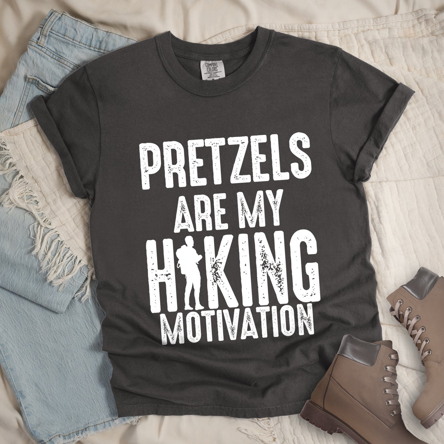Pepper (Dark Gray) t-shirt with white distressed text reading "PRETZELS ARE MY HIKING MOTIVATION". "PRETZELS" appears at the top, "ARE MY" in the middle, and "HIKING" in large letters with a hiker silhouette integrated into the letter "I". "MOTIVATION" appears at the bottom. Features a classic crew neck and casual rolled sleeves, styled alongside light wash denim and brown hiking boots.