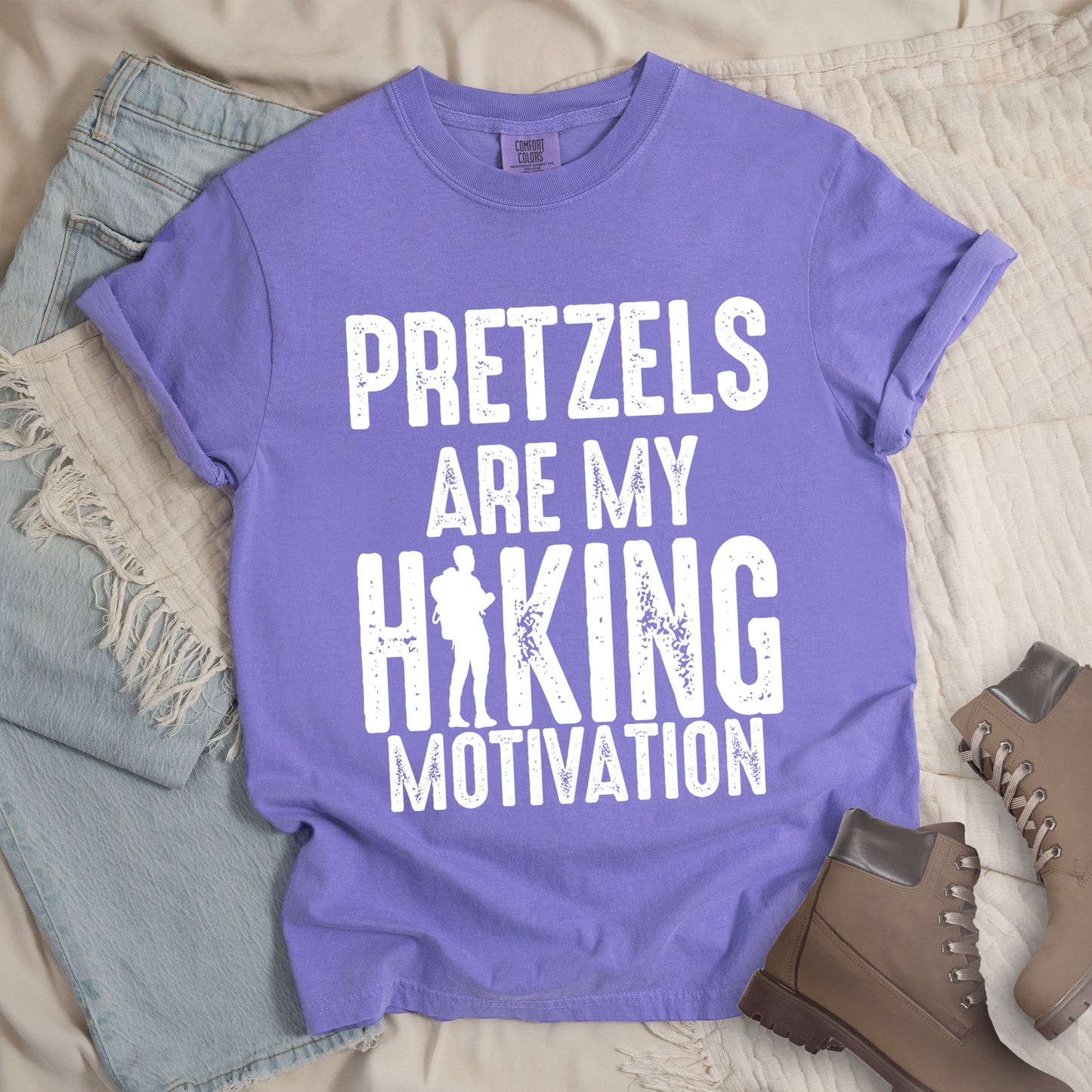 Violet t-shirt with white distressed text reading "PRETZELS ARE MY HIKING MOTIVATION". "PRETZELS" appears at the top, "ARE MY" in the middle, and "HIKING" in large letters with a hiker silhouette integrated into the letter "I". "MOTIVATION" appears at the bottom. Features a classic crew neck and casual rolled sleeves, styled alongside light wash denim and brown hiking boots.