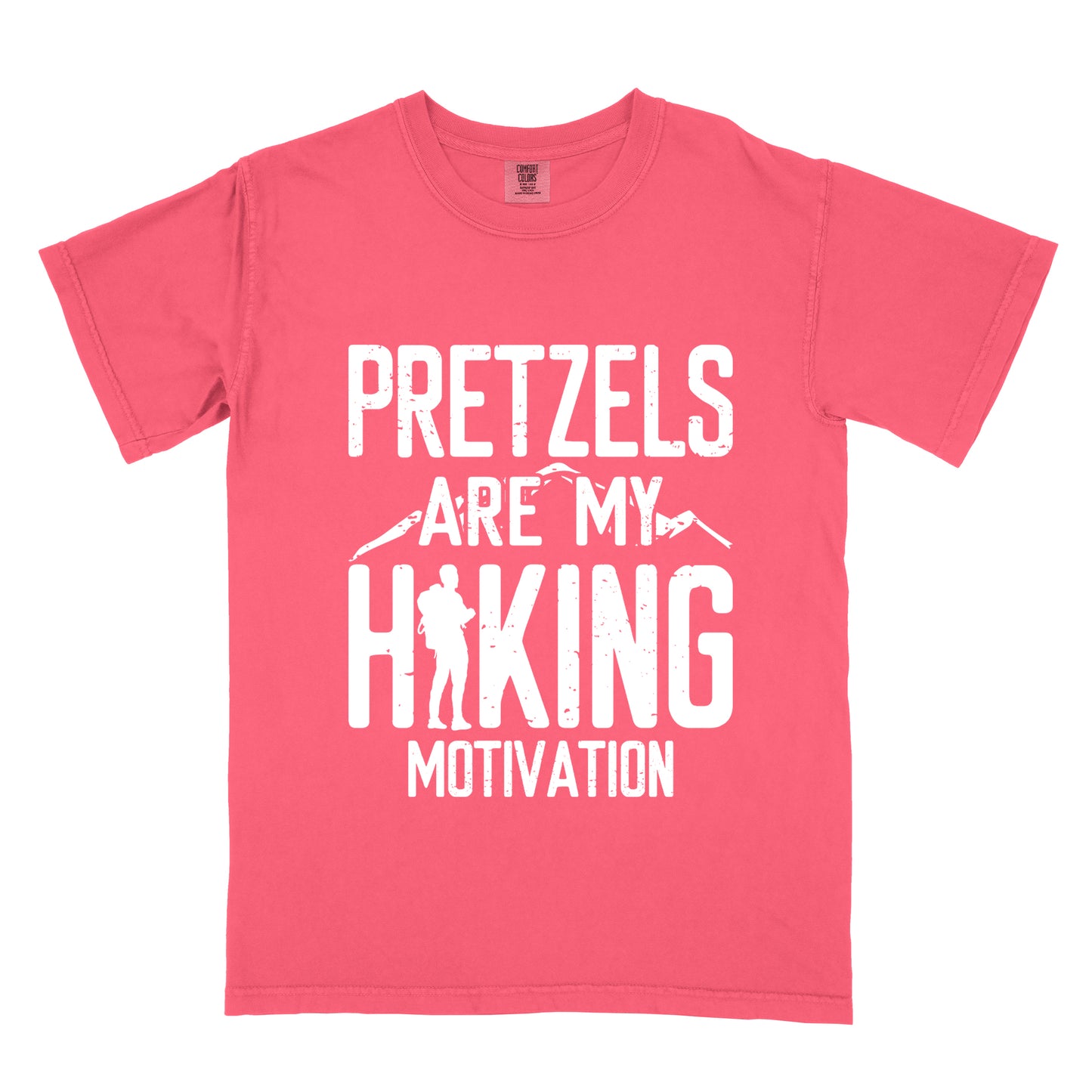 Watermelon (Light Coral) t-shirt with white distressed text reading "PRETZELS ARE MY HIKING MOTIVATION". "PRETZELS" appears at the top, "ARE MY" in the middle with small mountain silhouettes behind it, and "HIKING" in large letters with a hiker silhouette integrated into the letter "H". "MOTIVATION" appears at the bottom.