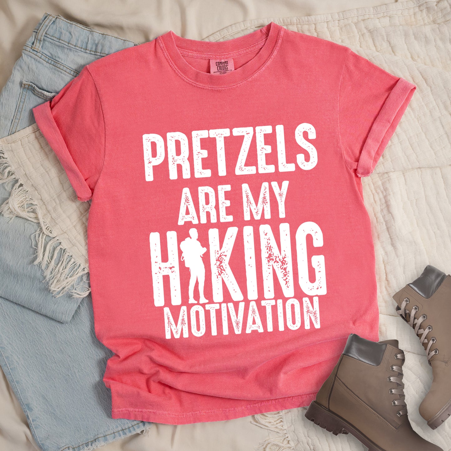 Watermelon (Light Coral) t-shirt with white distressed text reading "PRETZELS ARE MY HIKING MOTIVATION". "PRETZELS" appears at the top, "ARE MY" in the middle, and "HIKING" in large letters with a hiker silhouette integrated into the letter "I". "MOTIVATION" appears at the bottom. Features a classic crew neck and casual rolled sleeves, styled alongside light wash denim and brown hiking boots.