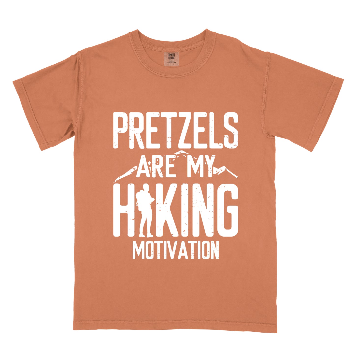 Yam (Brown) t-shirt with white distressed text reading "PRETZELS ARE MY HIKING MOTIVATION". "PRETZELS" appears at the top, "ARE MY" in the middle with small mountain silhouettes behind it, and "HIKING" in large letters with a hiker silhouette integrated into the letter "H". "MOTIVATION" appears at the bottom.