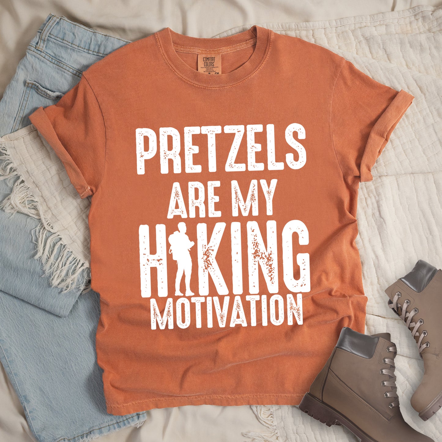 Yam (Brown) t-shirt with white distressed text reading "PRETZELS ARE MY HIKING MOTIVATION". "PRETZELS" appears at the top, "ARE MY" in the middle, and "HIKING" in large letters with a hiker silhouette integrated into the letter "I". "MOTIVATION" appears at the bottom. Features a classic crew neck and casual rolled sleeves, styled alongside light wash denim and brown hiking boots.