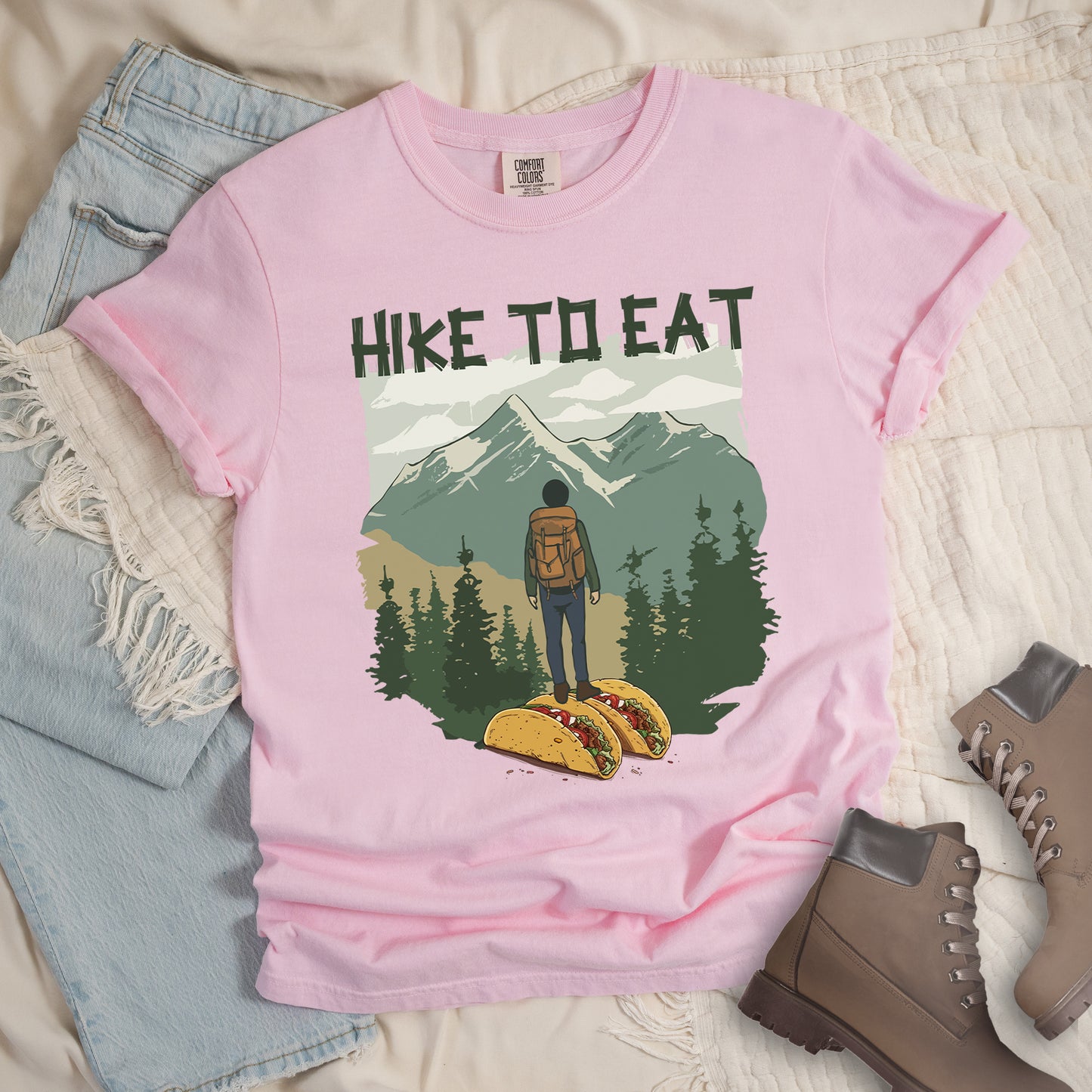 Light Pink (Blossom) t-shirt with "HIKE TO EAT" printed at the top in dark green text. The main graphic shows a hiker with backpack standing on two tacos, facing a scenic mountain landscape with pine trees in the foreground and snow-capped peaks in the background. The shirt features a classic crew neck and casual rolled sleeves, styled alongside light wash denim and brown hiking boots.