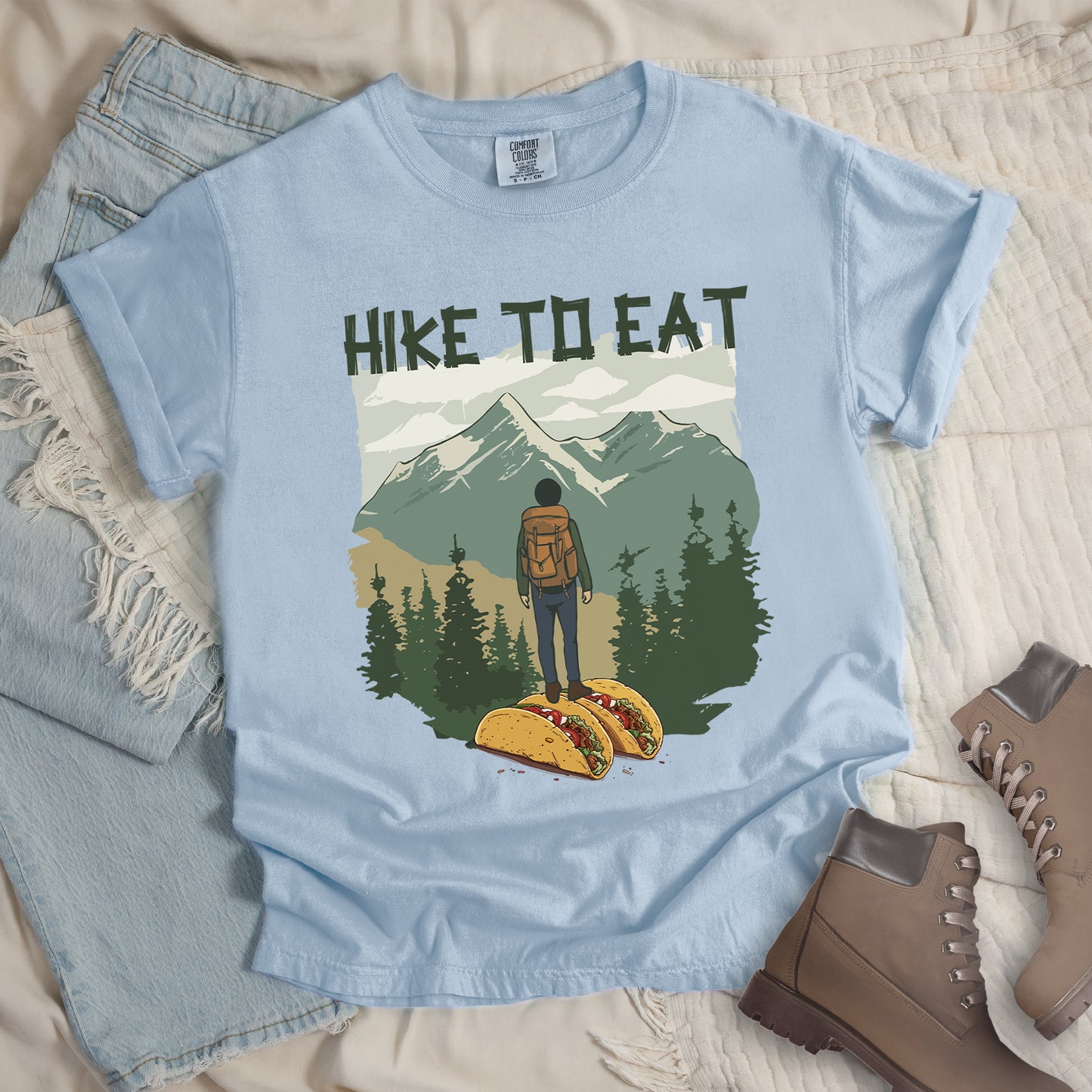 Light Blue (Chambray) t-shirt with "HIKE TO EAT" printed at the top in dark green text. The main graphic shows a hiker with backpack standing on two tacos, facing a scenic mountain landscape with pine trees in the foreground and snow-capped peaks in the background. The shirt features a classic crew neck and casual rolled sleeves, styled alongside light wash denim and brown hiking boots.