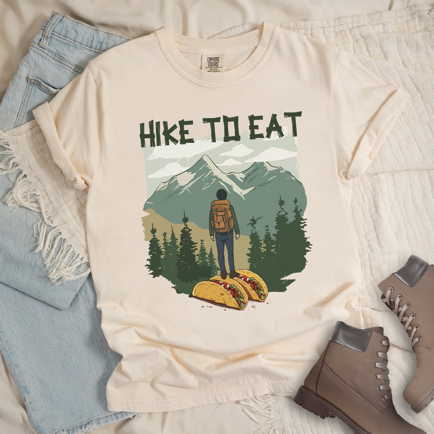 Cream-colored (ivory) t-shirt with "HIKE TO EAT" printed at the top in dark green text. The main graphic shows a hiker with backpack standing on two tacos, facing a scenic mountain landscape with pine trees in the foreground and snow-capped peaks in the background. The shirt features a classic crew neck and casual rolled sleeves, styled alongside light wash denim and brown hiking boots.