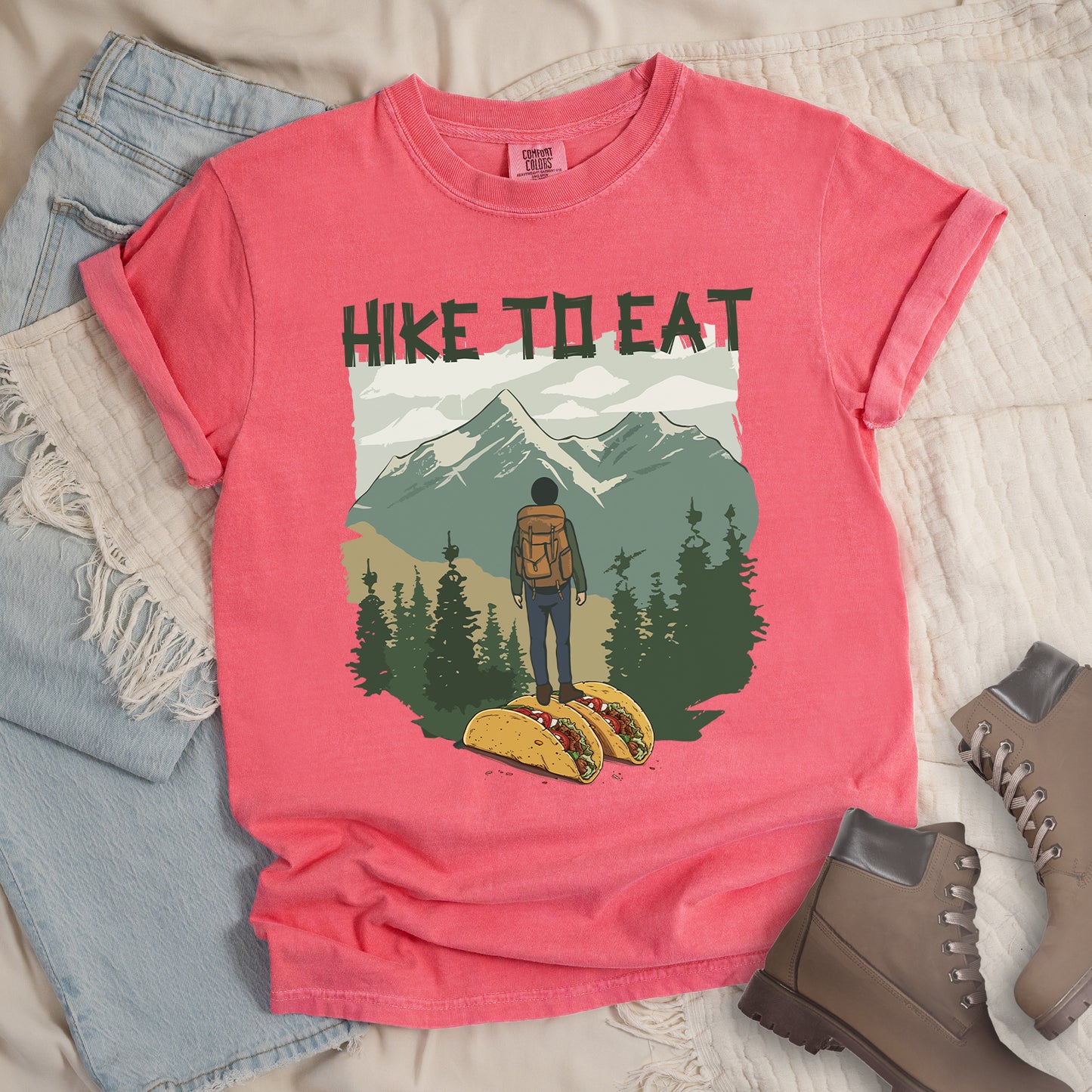 Watermelon (Light coral) t-shirt with "HIKE TO EAT" printed at the top in dark green text. The main graphic shows a hiker with backpack standing on two tacos, facing a scenic mountain landscape with pine trees in the foreground and snow-capped peaks in the background. The shirt features a classic crew neck and casual rolled sleeves, styled alongside light wash denim and brown hiking boots.