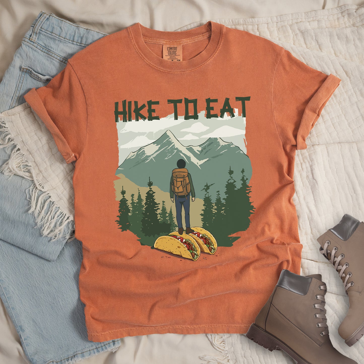 Yam colored (Brown) t-shirt with "HIKE TO EAT" printed at the top in dark green text. The main graphic shows a hiker with backpack standing on two tacos, facing a scenic mountain landscape with pine trees in the foreground and snow-capped peaks in the background. The shirt features a classic crew neck and casual rolled sleeves, styled alongside light wash denim and brown hiking boots.