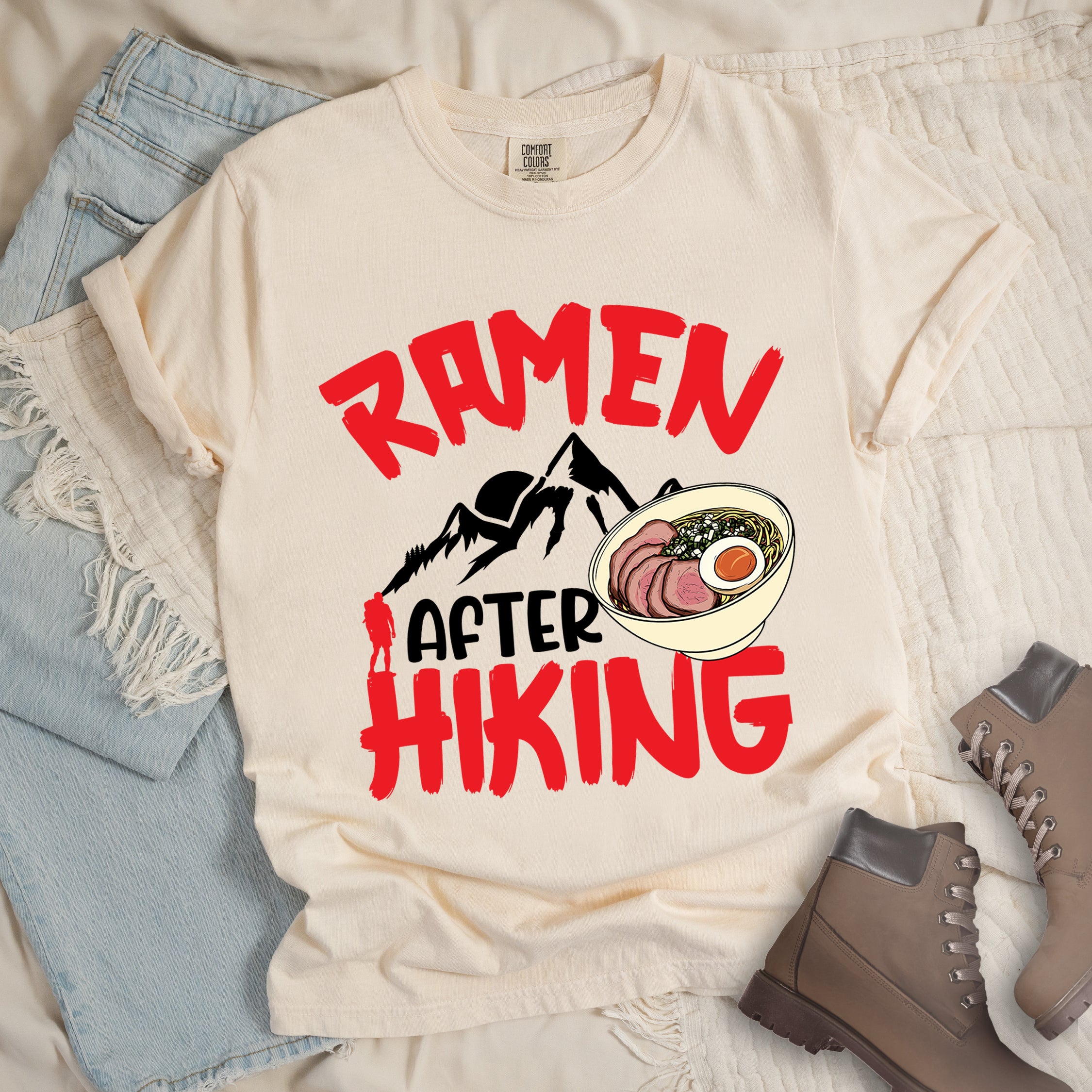 Ivory (Cream White) t-shirt with red text reading "RAMEN AFTER HIKING". "RAMEN" appears in bold red letters at the top, "AFTER" in black in the middle, and "HIKING" in bold red at the bottom. Features black mountain silhouettes in the center with a red hiker figure, and a detailed illustration of a ramen bowl with sliced meat, egg, and green onions on the right. The design connects outdoor activities with food rewards.