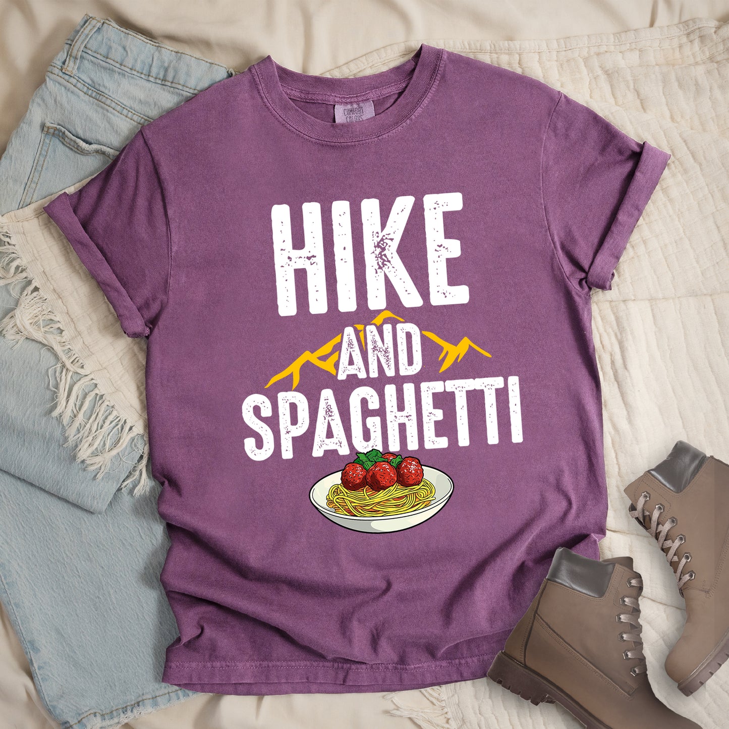 Berry (Dark Violet) t-shirt with white distressed text reading "HIKE AND SPAGHETTI". "HIKE" appears in large white letters at the top, "AND" in white over yellow mountain silhouettes in the middle, and "SPAGHETTI" in large white letters at the bottom. Features a detailed illustration of a white plate with spaghetti, meatballs, and herbs at the bottom of the design.Features a classic crew neck and casual rolled sleeves, styled alongside light wash denim and brown hiking boots.