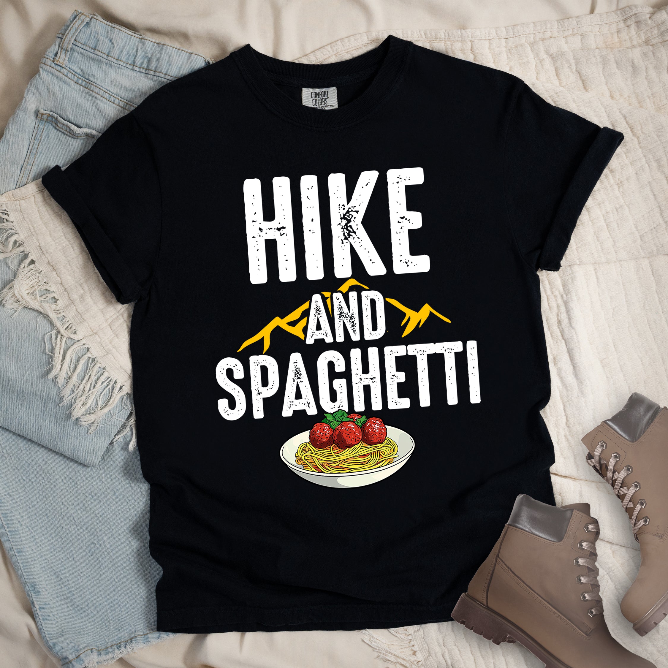 Black t-shirt with white distressed text reading "HIKE AND SPAGHETTI". "HIKE" appears in large white letters at the top, "AND" in white over yellow mountain silhouettes in the middle, and "SPAGHETTI" in large white letters at the bottom. Features a detailed illustration of a white plate with spaghetti, meatballs, and herbs at the bottom of the design.Features a classic crew neck and casual rolled sleeves, styled alongside light wash denim and brown hiking boots.
