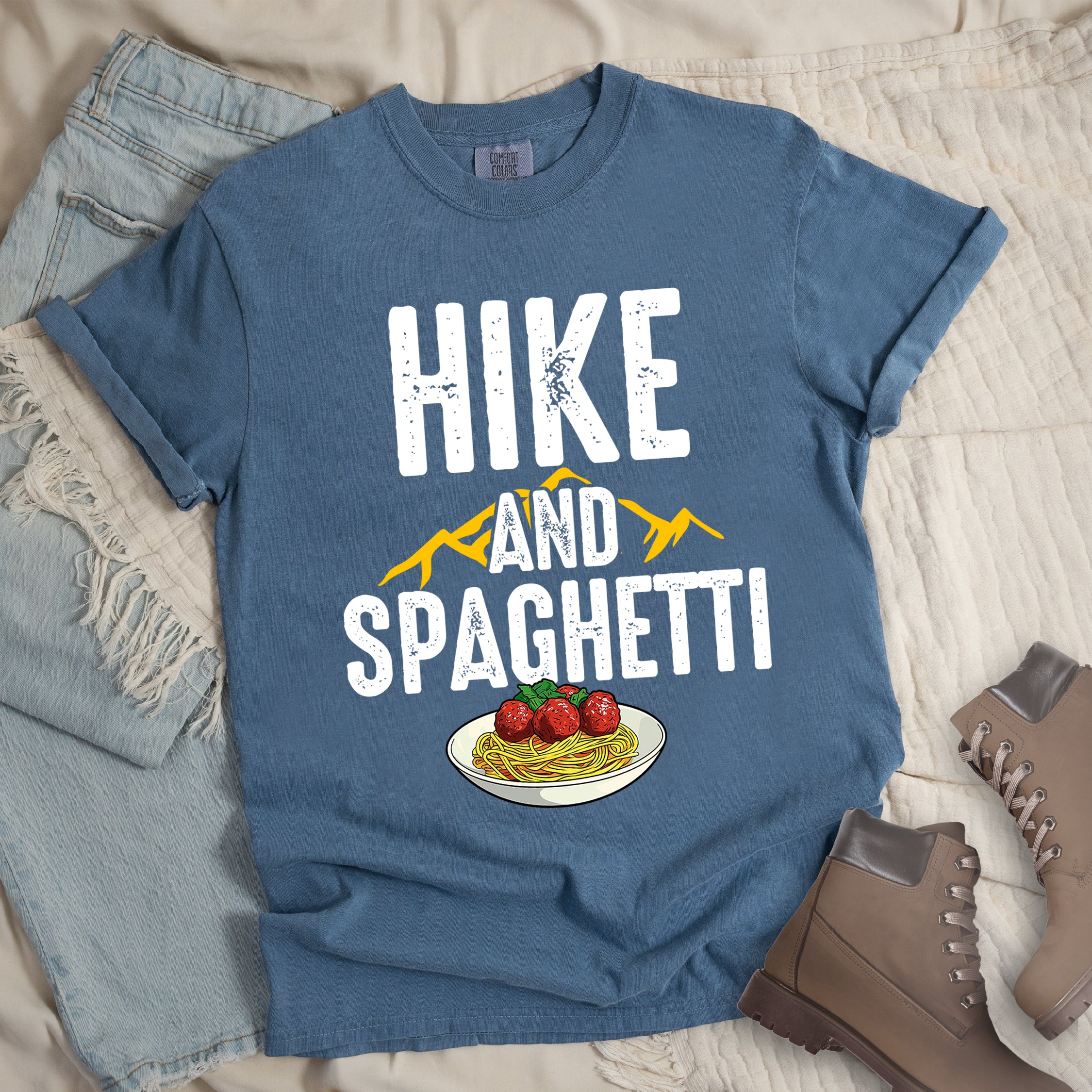 Blue Jean (Denim Blue) t-shirt with white distressed text reading "HIKE AND SPAGHETTI". "HIKE" appears in large white letters at the top, "AND" in white over yellow mountain silhouettes in the middle, and "SPAGHETTI" in large white letters at the bottom. Features a detailed illustration of a white plate with spaghetti, meatballs, and herbs at the bottom of the design.Features a classic crew neck and casual rolled sleeves, styled alongside light wash denim and brown hiking boots.