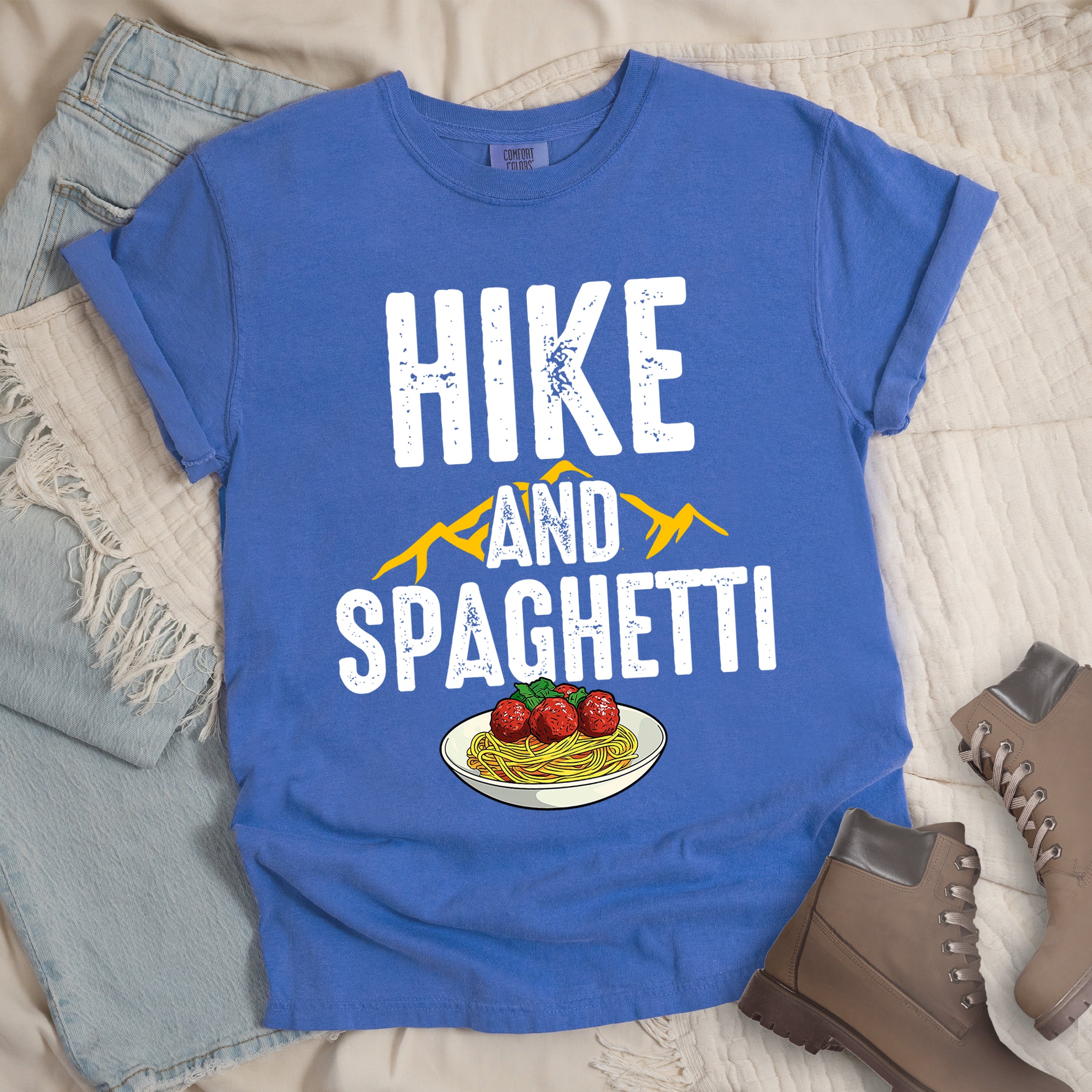 Flo Blue (Bright Blue) t-shirt with white distressed text reading "HIKE AND SPAGHETTI". "HIKE" appears in large white letters at the top, "AND" in white over yellow mountain silhouettes in the middle, and "SPAGHETTI" in large white letters at the bottom. Features a detailed illustration of a white plate with spaghetti, meatballs, and herbs at the bottom of the design.Features a classic crew neck and casual rolled sleeves, styled alongside light wash denim and brown hiking boots.