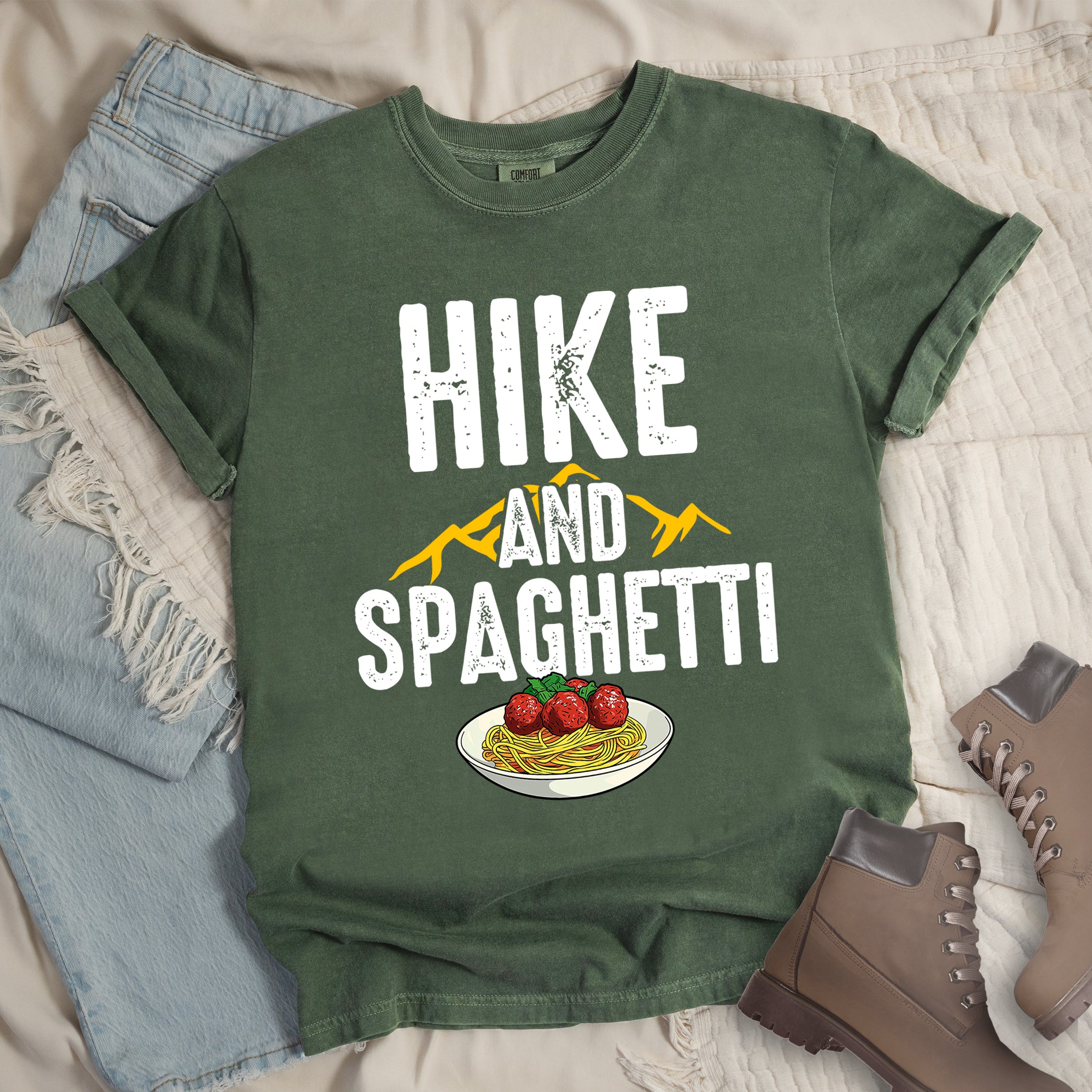 Moss (Dark Green) t-shirt with white distressed text reading "HIKE AND SPAGHETTI". "HIKE" appears in large white letters at the top, "AND" in white over yellow mountain silhouettes in the middle, and "SPAGHETTI" in large white letters at the bottom. Features a detailed illustration of a white plate with spaghetti, meatballs, and herbs at the bottom of the design.Features a classic crew neck and casual rolled sleeves, styled alongside light wash denim and brown hiking boots.