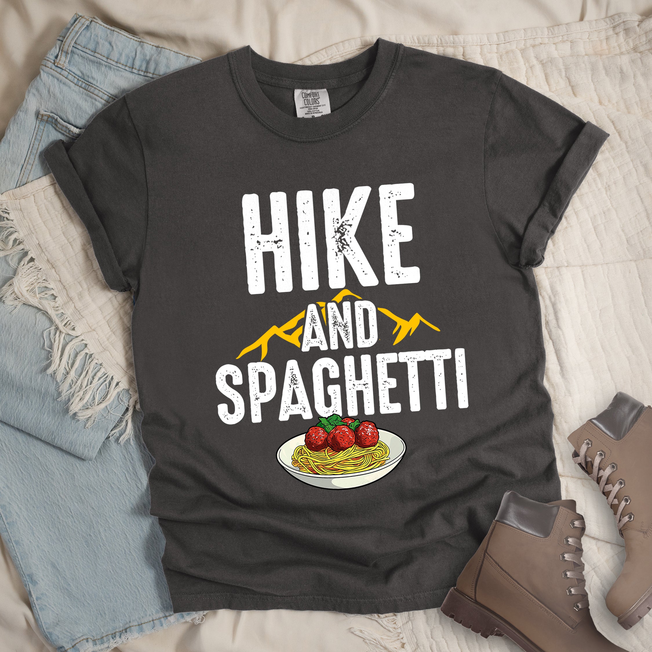 Pepper (Dark Gray) t-shirt with white distressed text reading "HIKE AND SPAGHETTI". "HIKE" appears in large white letters at the top, "AND" in white over yellow mountain silhouettes in the middle, and "SPAGHETTI" in large white letters at the bottom. Features a detailed illustration of a white plate with spaghetti, meatballs, and herbs at the bottom of the design.Features a classic crew neck and casual rolled sleeves, styled alongside light wash denim and brown hiking boots.
