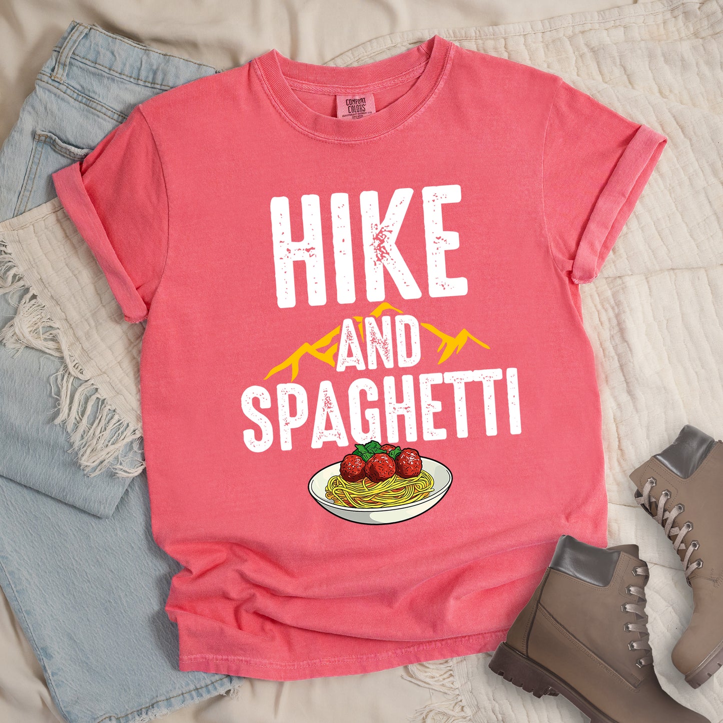Watermelon (Light Coral) t-shirt with white distressed text reading "HIKE AND SPAGHETTI". "HIKE" appears in large white letters at the top, "AND" in white over yellow mountain silhouettes in the middle, and "SPAGHETTI" in large white letters at the bottom. Features a detailed illustration of a white plate with spaghetti, meatballs, and herbs at the bottom of the design.Features a classic crew neck and casual rolled sleeves, styled alongside light wash denim and brown hiking boots.