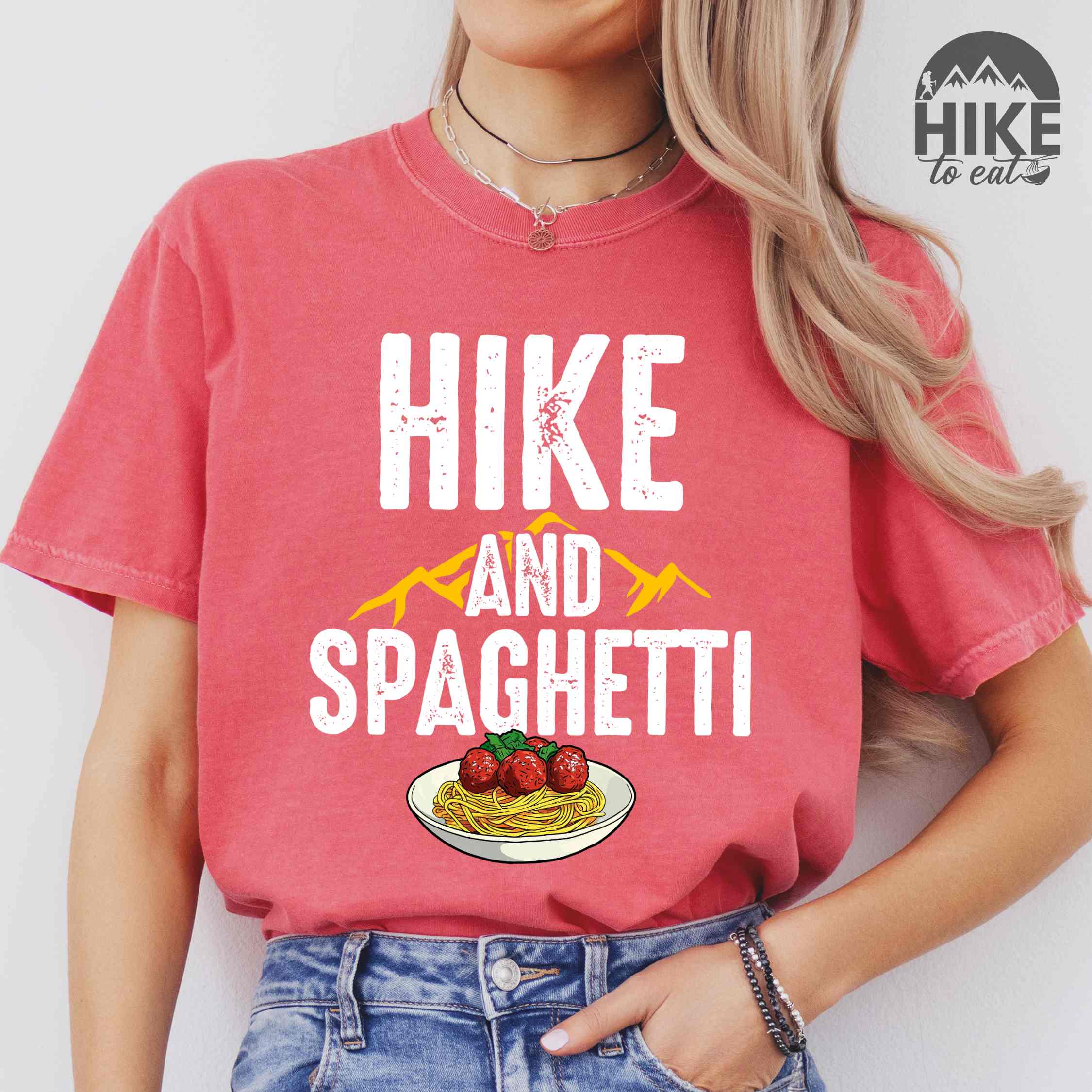 Woman wearing watermelon "Hike and Spaghetti" colored vintage tee with bold typography, birds, mountains, illustrated plate of spaghetti and meatballs, and backpacker for pasta lovers.