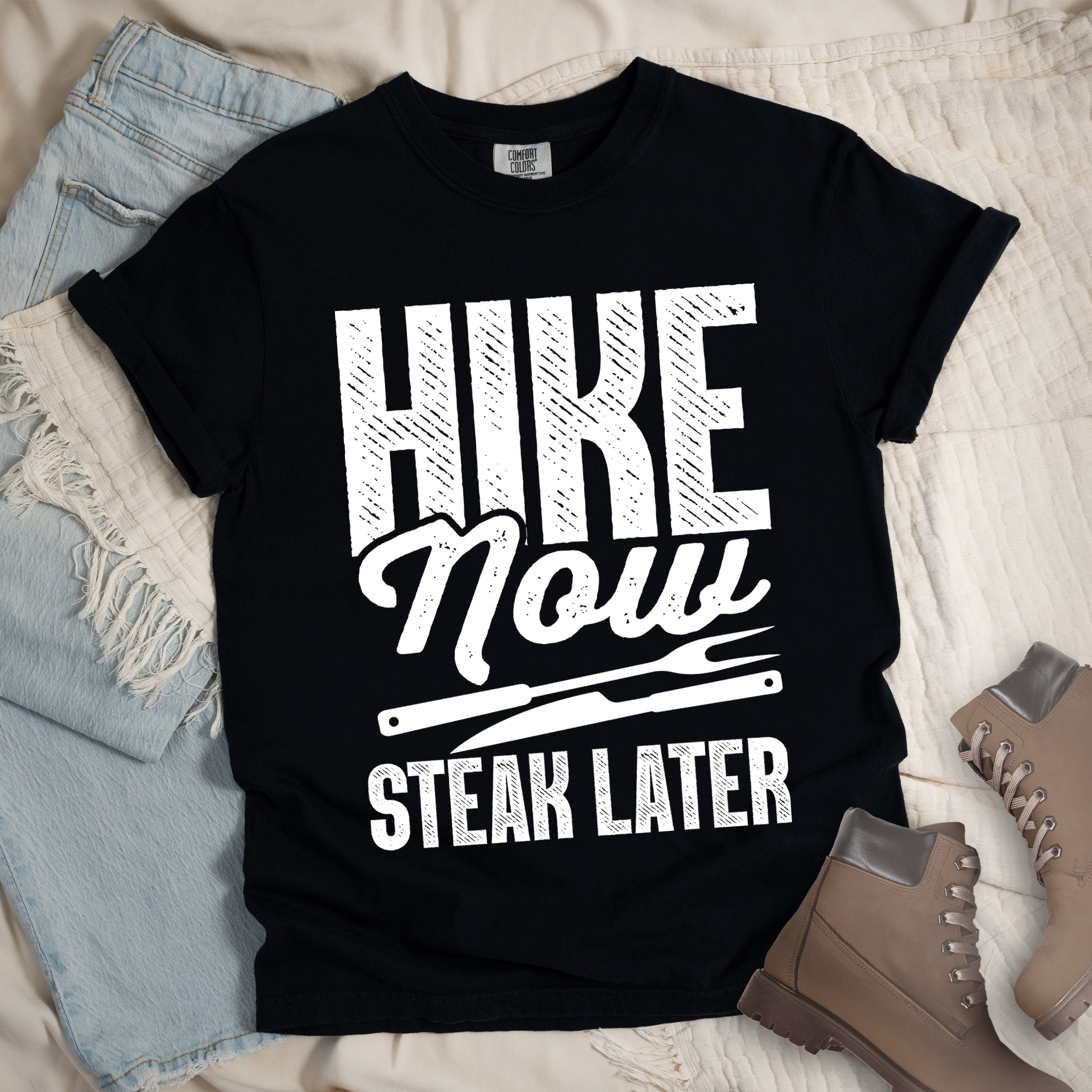 Black t-shirt with white distressed text reading "HIKE Now STEAK LATER". "HIKE" appears in large bold block letters at the top, "Now" in stylish script in the middle, and "STEAK LATER" in block letters at the bottom. Between the middle and bottom text is a simple illustration of a steak knife. The design emphasizes the reward of enjoying steak after completing a hiking activity. Features a classic crew neck and casual rolled sleeves, styled alongside light wash denim and brown hiking boots.