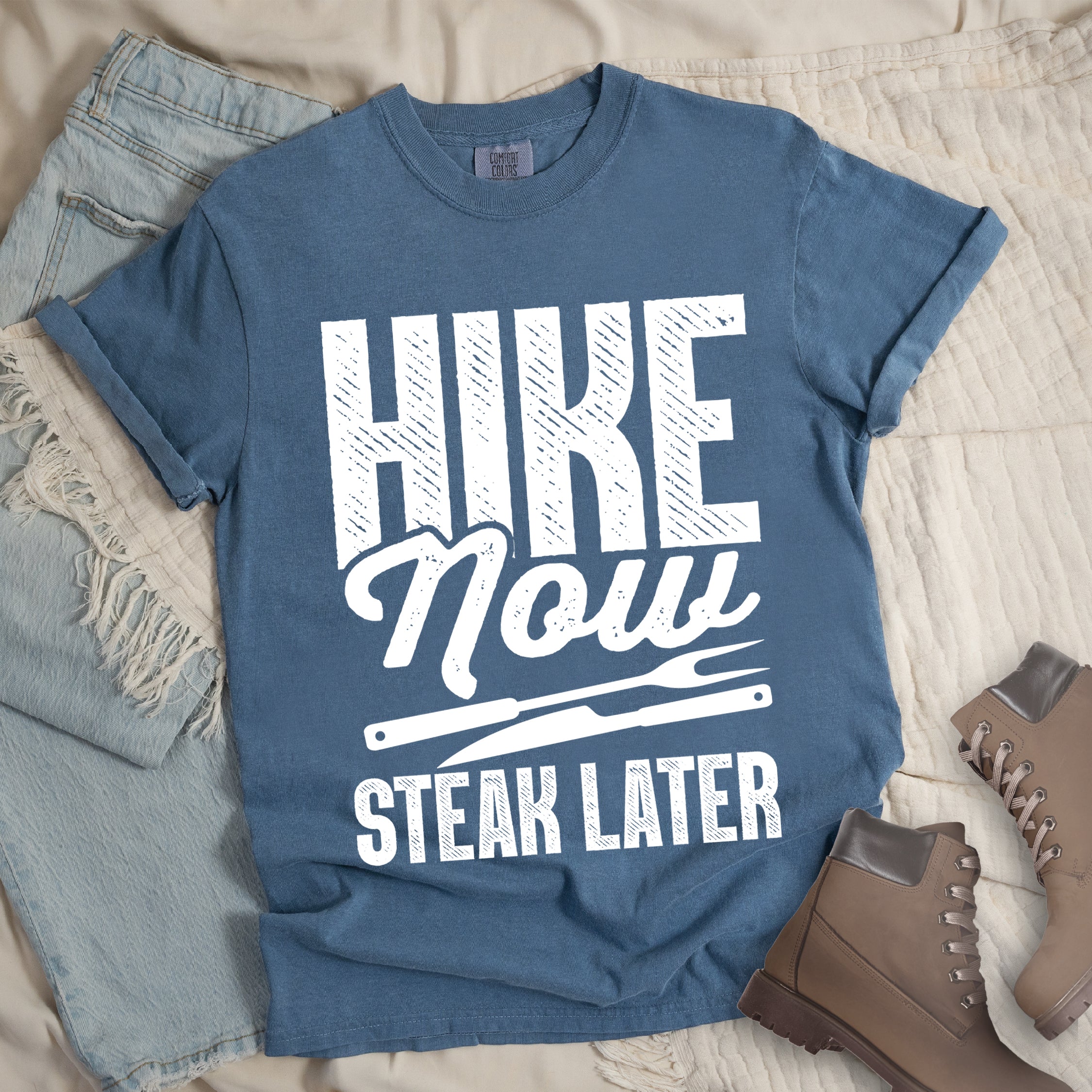 Blue Jean (Denim Blue) t-shirt with white distressed text reading "HIKE Now STEAK LATER". "HIKE" appears in large bold block letters at the top, "Now" in stylish script in the middle, and "STEAK LATER" in block letters at the bottom. Between the middle and bottom text is a simple illustration of a steak knife. The design emphasizes the reward of enjoying steak after completing a hiking activity. Features a classic crew neck and casual rolled sleeves, styled alongside light wash denim and brown hiking boots.