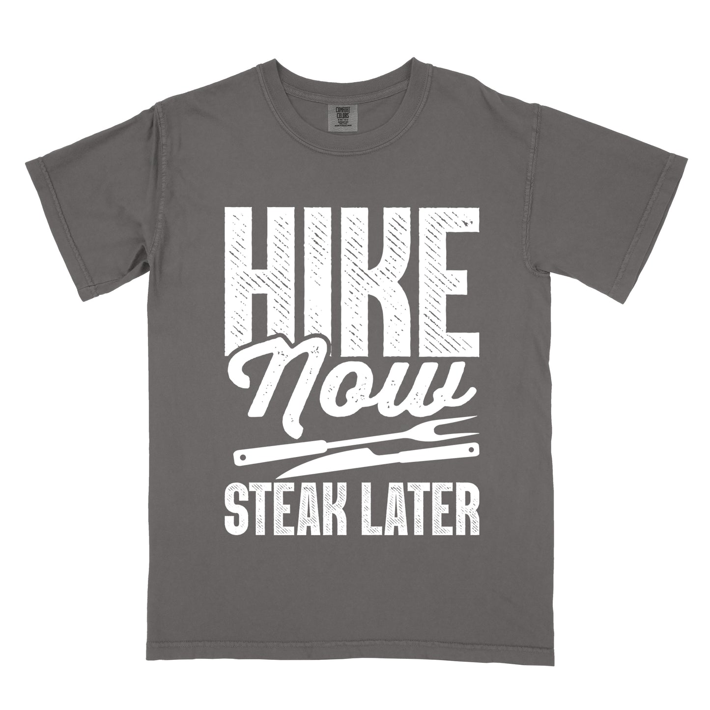 Pepper (Dark Gray) t-shirt with white distressed text reading "HIKE Now STEAK LATER". "HIKE" appears in large bold block letters at the top, "Now" in stylish script in the middle, and "STEAK LATER" in block letters at the bottom. Between the middle and bottom text is a simple illustration of a steak knife. The design emphasizes the reward of enjoying steak after completing a hiking activity.