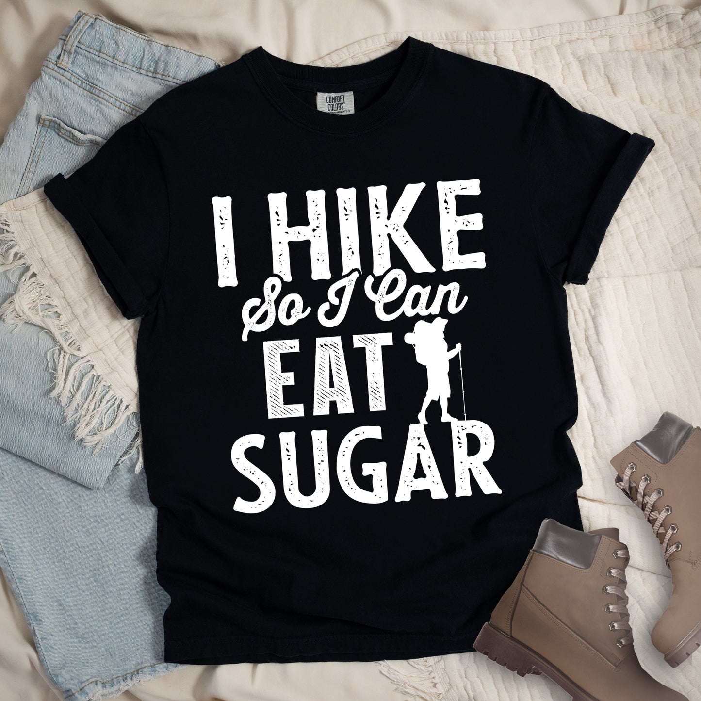 Black t-shirt with white distressed text reading "I HIKE So I Can EAT SUGAR". "I HIKE" appears in large block letters at the top, "So I Can" in script in the middle, and "EAT SUGAR" in block letters at the bottom. A small silhouette of a hiker with backpack and walking stick is positioned next to the word "EAT" and above "SUGAR". Features a classic crew neck and casual rolled sleeves, styled alongside light wash denim and brown hiking boots.
