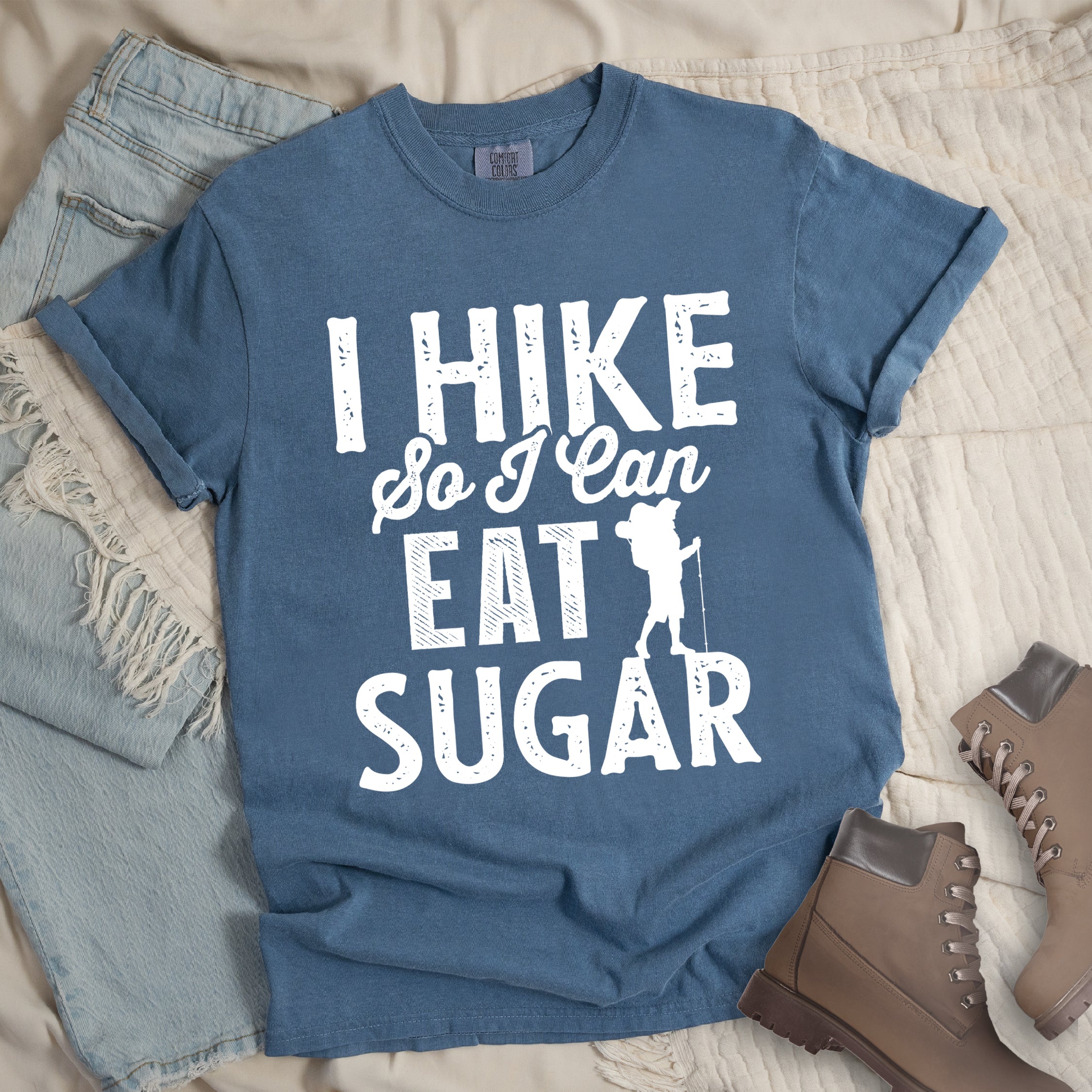 Blue Jean (Denim Blue) t-shirt with white distressed text reading "I HIKE So I Can EAT SUGAR". "I HIKE" appears in large block letters at the top, "So I Can" in script in the middle, and "EAT SUGAR" in block letters at the bottom. A small silhouette of a hiker with backpack and walking stick is positioned next to the word "EAT" and above "SUGAR". Features a classic crew neck and casual rolled sleeves, styled alongside light wash denim and brown hiking boots.