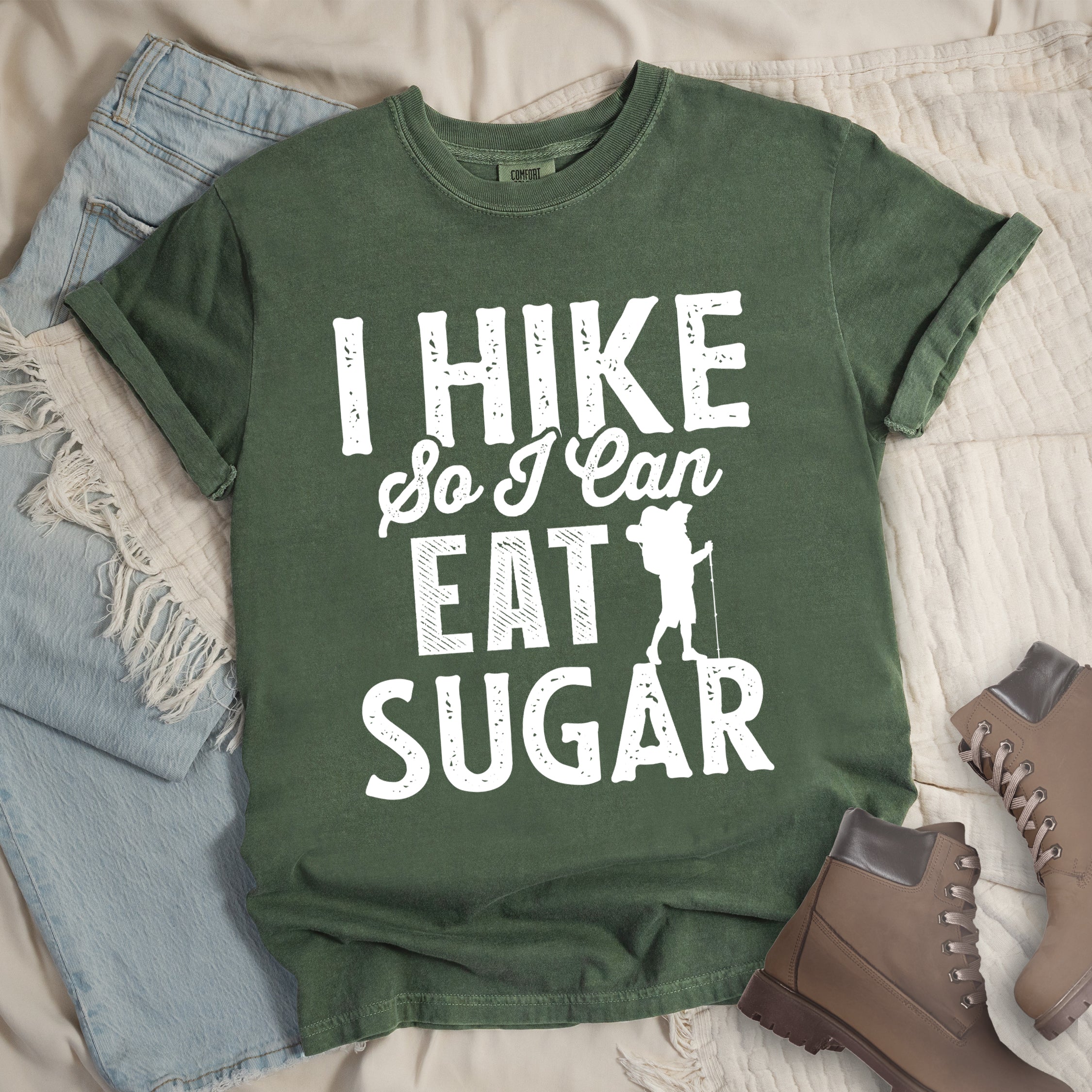 Moss (Dark Green) t-shirt with white distressed text reading "I HIKE So I Can EAT SUGAR". "I HIKE" appears in large block letters at the top, "So I Can" in script in the middle, and "EAT SUGAR" in block letters at the bottom. A small silhouette of a hiker with backpack and walking stick is positioned next to the word "EAT" and above "SUGAR". Features a classic crew neck and casual rolled sleeves, styled alongside light wash denim and brown hiking boots.