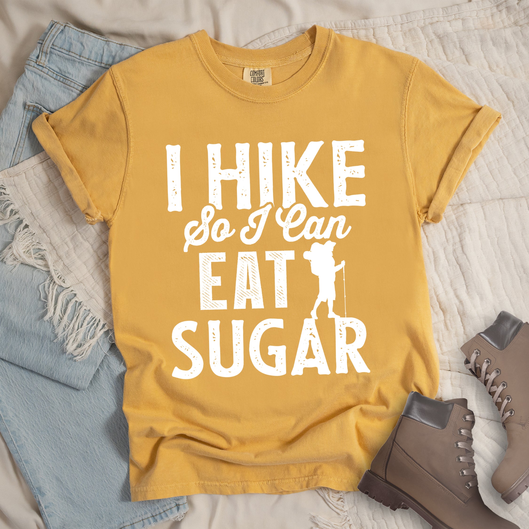 Mustard (Yellow) t-shirt with white distressed text reading "I HIKE So I Can EAT SUGAR". "I HIKE" appears in large block letters at the top, "So I Can" in script in the middle, and "EAT SUGAR" in block letters at the bottom. A small silhouette of a hiker with backpack and walking stick is positioned next to the word "EAT" and above "SUGAR". Features a classic crew neck and casual rolled sleeves, styled alongside light wash denim and brown hiking boots.
