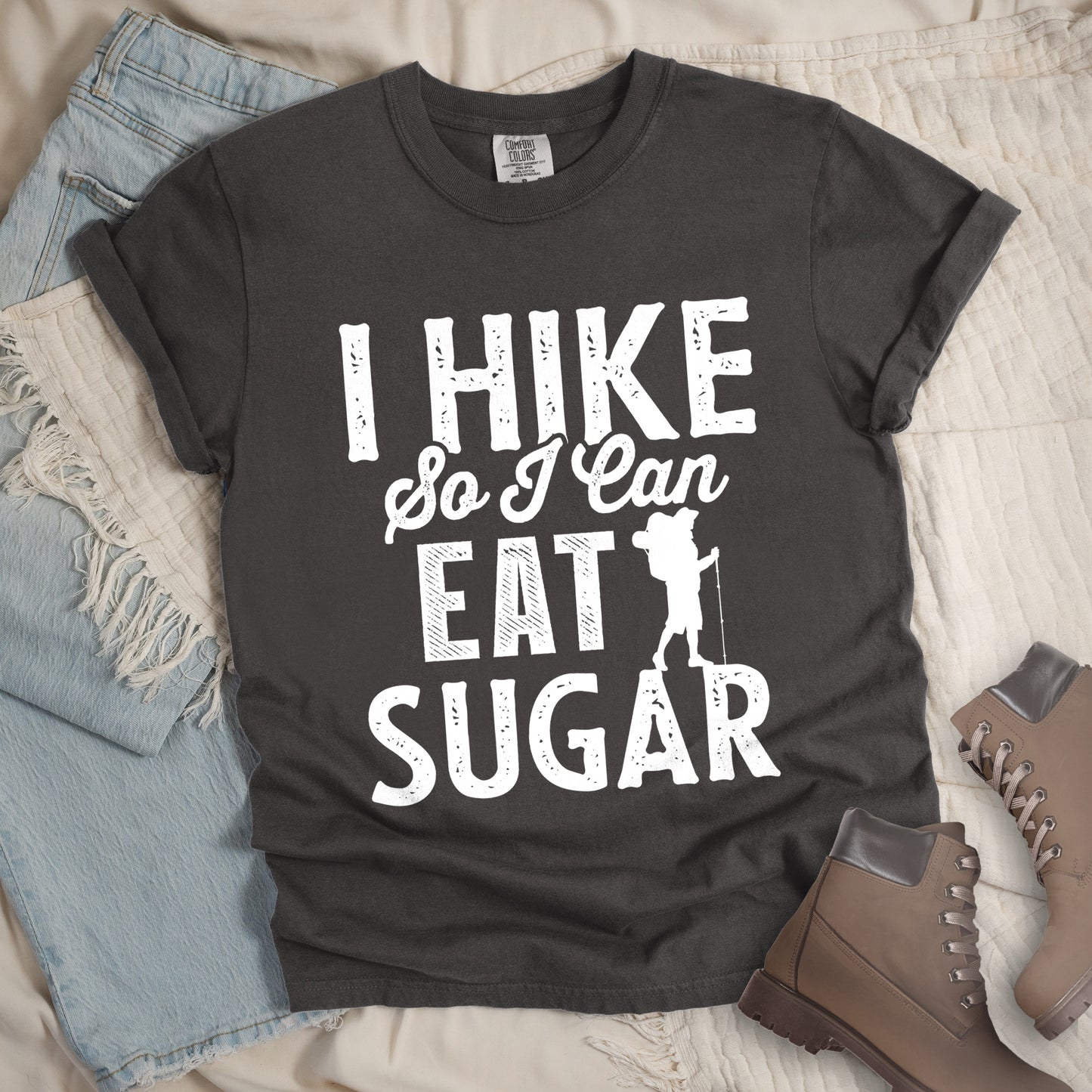 Pepper (Dark Gray) t-shirt with white distressed text reading "I HIKE So I Can EAT SUGAR". "I HIKE" appears in large block letters at the top, "So I Can" in script in the middle, and "EAT SUGAR" in block letters at the bottom. A small silhouette of a hiker with backpack and walking stick is positioned next to the word "EAT" and above "SUGAR". Features a classic crew neck and casual rolled sleeves, styled alongside light wash denim and brown hiking boots.