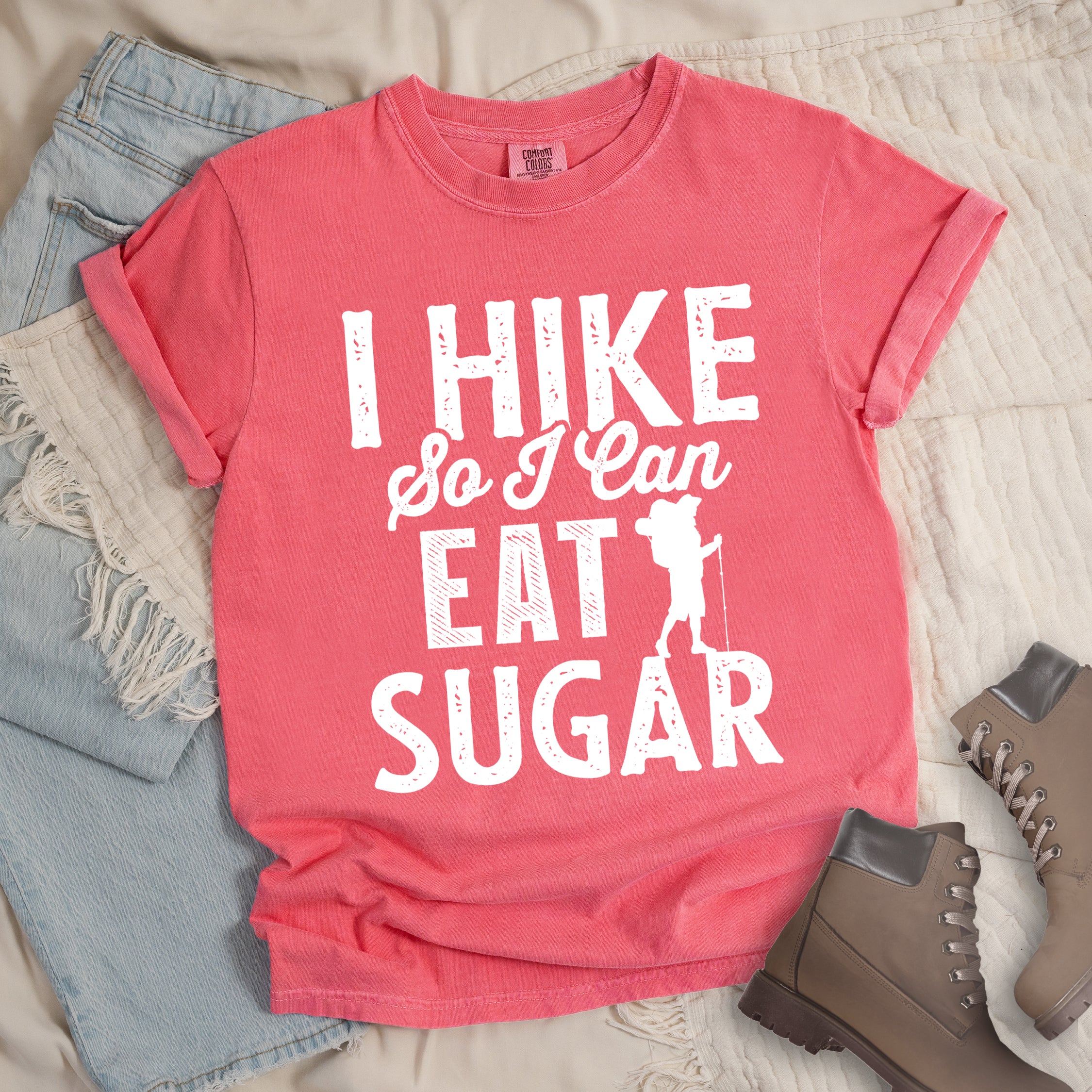 Watermelon (Light Coral) t-shirt with white distressed text reading "I HIKE So I Can EAT SUGAR". "I HIKE" appears in large block letters at the top, "So I Can" in script in the middle, and "EAT SUGAR" in block letters at the bottom. A small silhouette of a hiker with backpack and walking stick is positioned next to the word "EAT" and above "SUGAR". Features a classic crew neck and casual rolled sleeves, styled alongside light wash denim and brown hiking boots.
