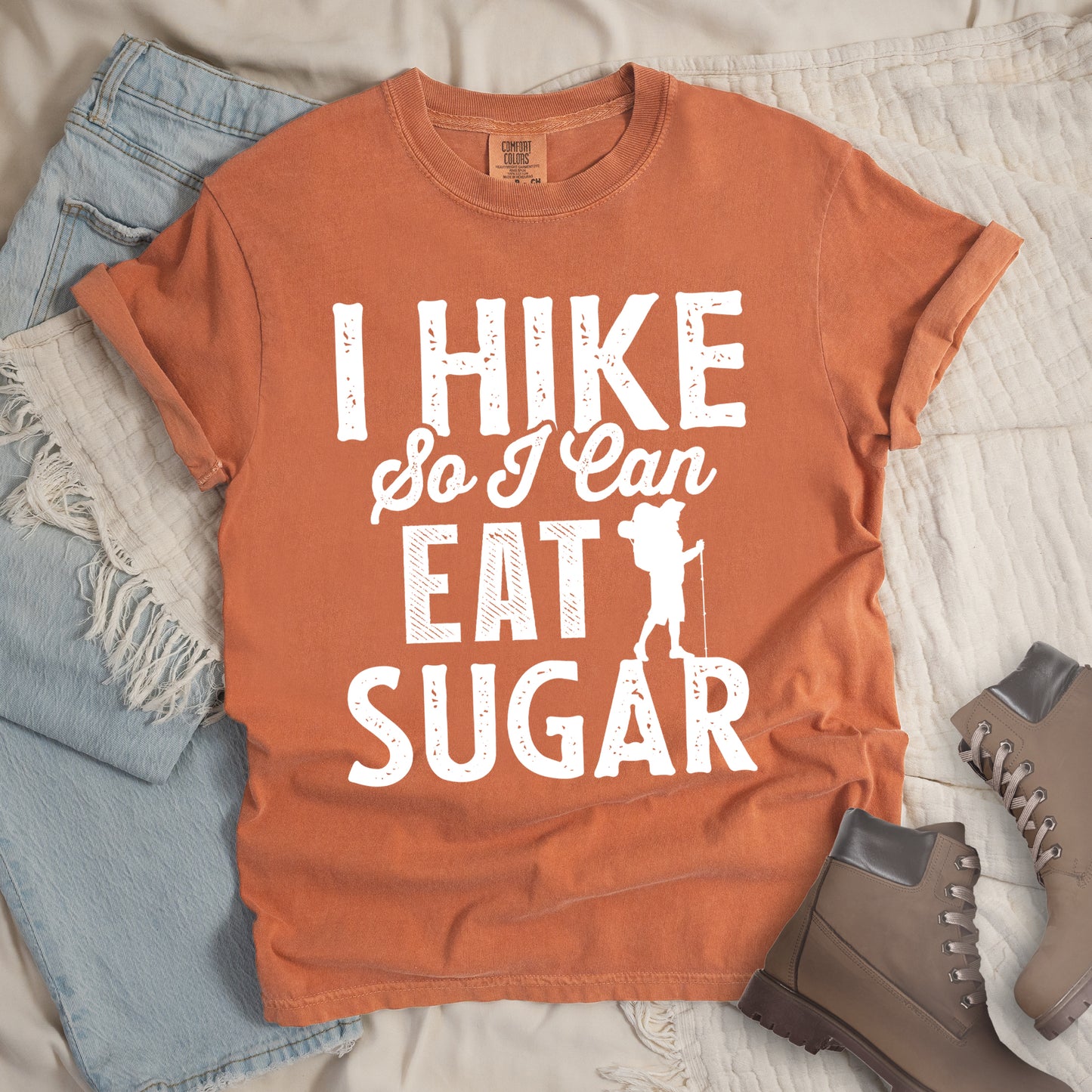 Yam (Brown) t-shirt with white distressed text reading "I HIKE So I Can EAT SUGAR". "I HIKE" appears in large block letters at the top, "So I Can" in script in the middle, and "EAT SUGAR" in block letters at the bottom. A small silhouette of a hiker with backpack and walking stick is positioned next to the word "EAT" and above "SUGAR". Features a classic crew neck and casual rolled sleeves, styled alongside light wash denim and brown hiking boots.