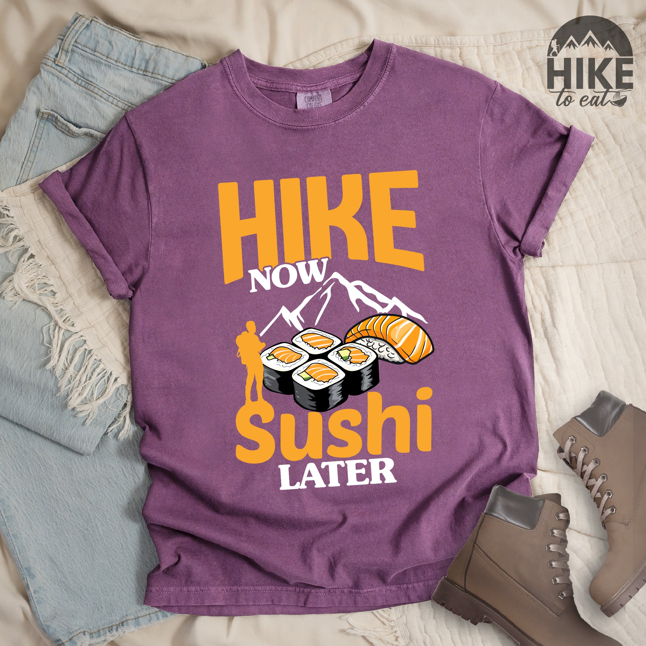 Berry (Dark Violet) t-shirt with orange and white text reading "HIKE NOW sushi LATER". "HIKE" appears in large orange letters at the top, "NOW" in white, "sushi" in orange at the bottom, and "LATER" in white. Features white mountain silhouettes in the center, detailed sushi illustrations including rolls and nigiri in orange/white, and an orange silhouette of a hiker with backpack on the left.