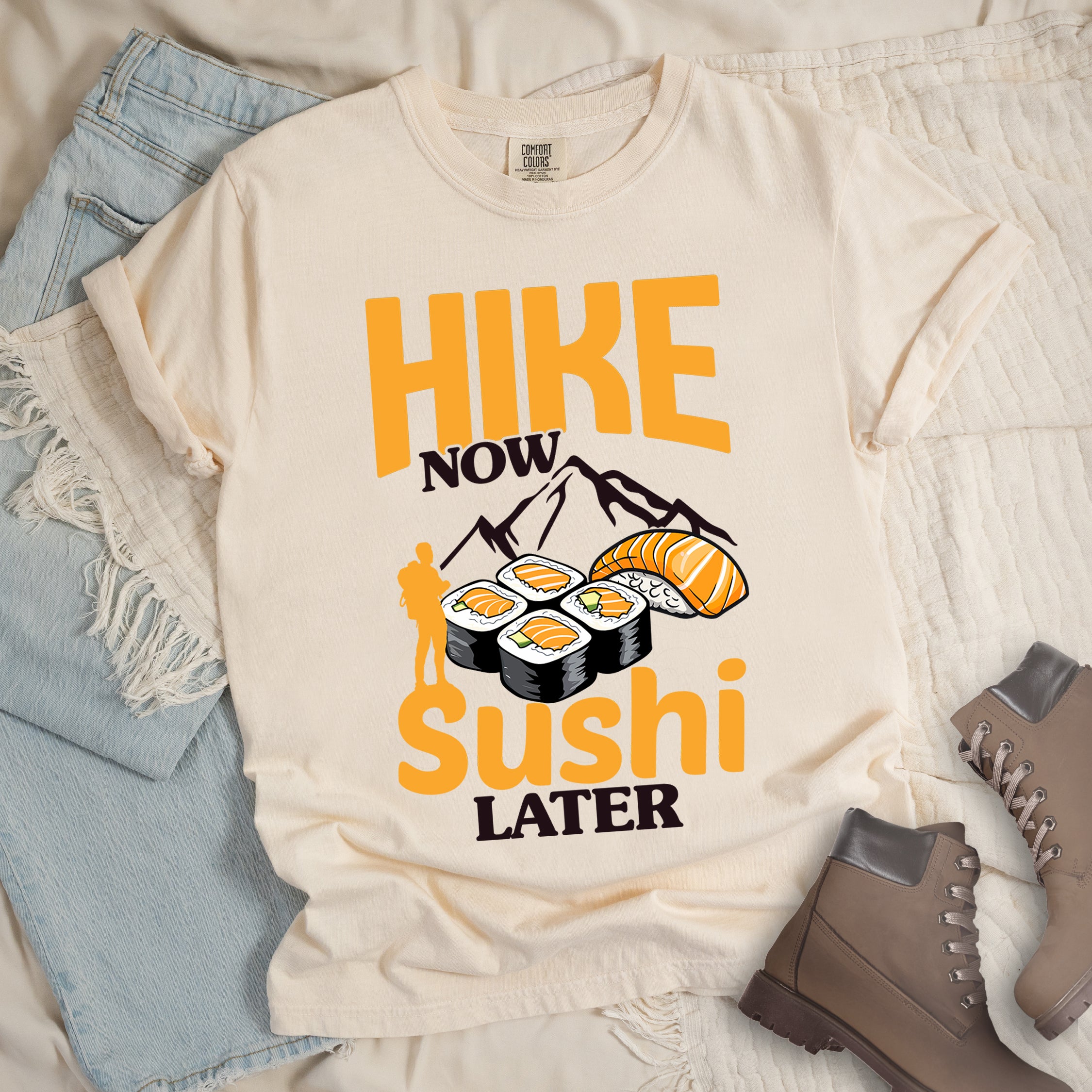Ivory (Cream White) t-shirt with orange and white text reading "HIKE NOW sushi LATER". "HIKE" appears in large orange letters at the top, "NOW" in white, "sushi" in orange at the bottom, and "LATER" in white. Features white mountain silhouettes in the center, detailed sushi illustrations including rolls and nigiri in orange/white, and an orange silhouette of a hiker with backpack on the left.