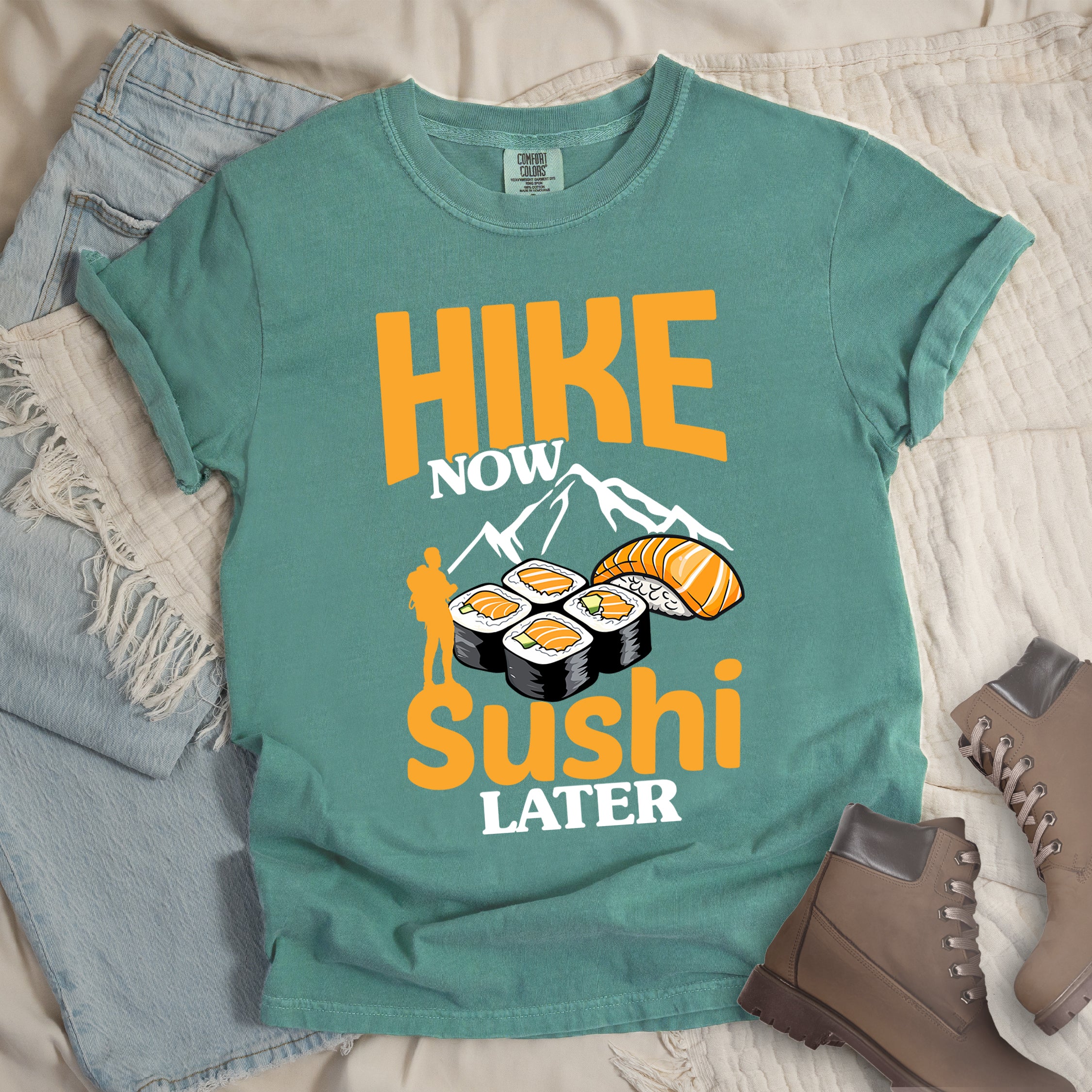 Light Green t-shirt with orange and white text reading "HIKE NOW sushi LATER". "HIKE" appears in large orange letters at the top, "NOW" in white, "sushi" in orange at the bottom, and "LATER" in white. Features white mountain silhouettes in the center, detailed sushi illustrations including rolls and nigiri in orange/white, and an orange silhouette of a hiker with backpack on the left.