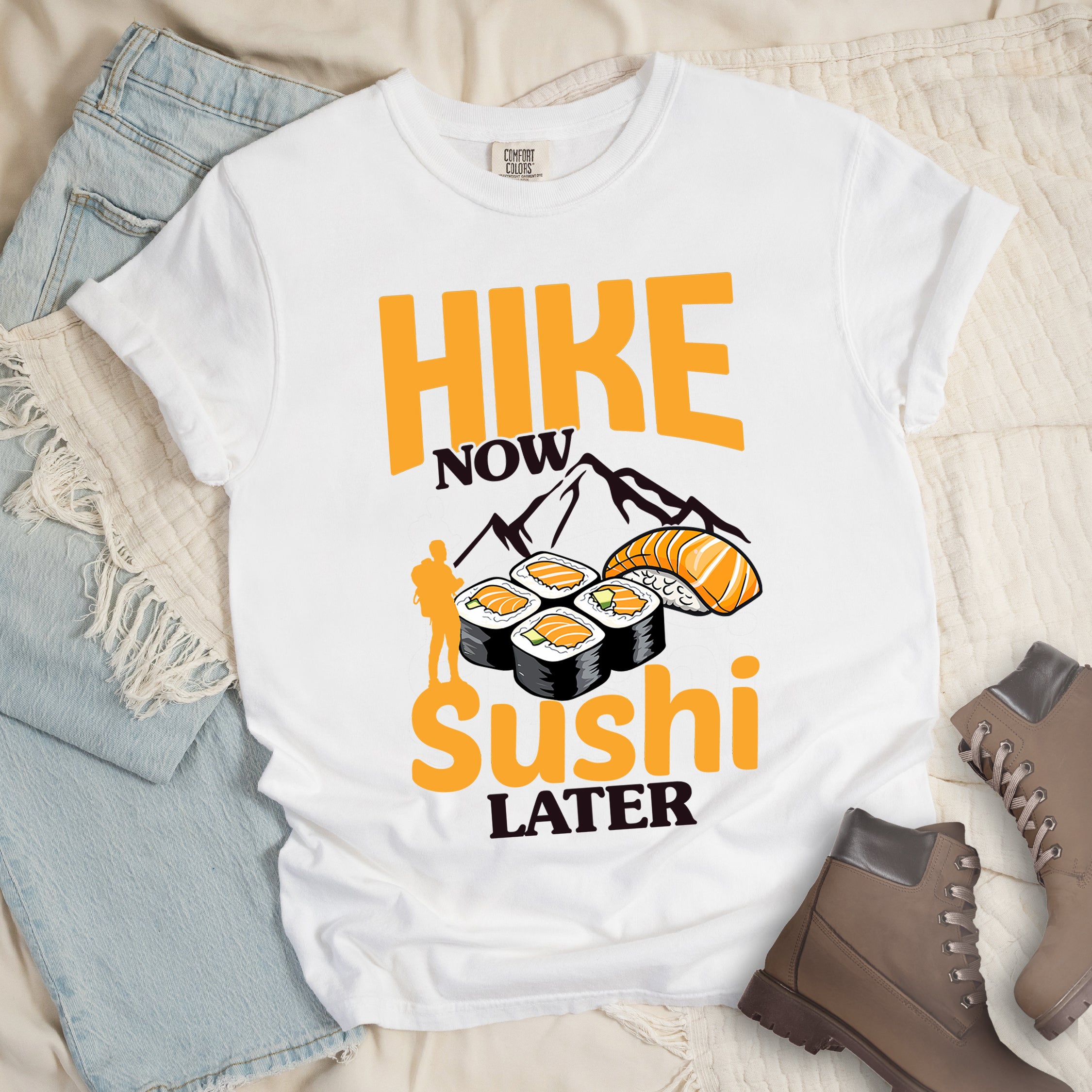 White t-shirt with orange and black text reading "HIKE NOW sushi LATER". "HIKE" appears in large orange letters at the top, "NOW" in black, "sushi" in orange at the bottom, and "LATER" in black. Features black mountain silhouettes in the center, detailed sushi illustrations including rolls and nigiri in orange/white, and an orange silhouette of a hiker with backpack on the left.