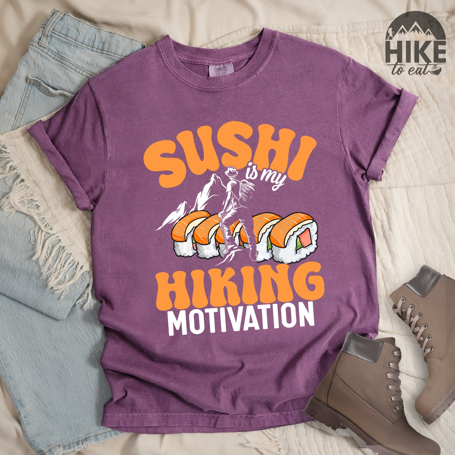 Berry (Dark Violet) t-shirt with orange and white text reading "SUSHI is my HIKING MOTIVATION". "SUSHI" appears in large orange letters at the top, "is my" in small white text, and "HIKING" in orange with "MOTIVATION" in white at the bottom. Features a humorous illustration of a hiker climbing over giant sushi rolls, with white mountain silhouettes in the background.