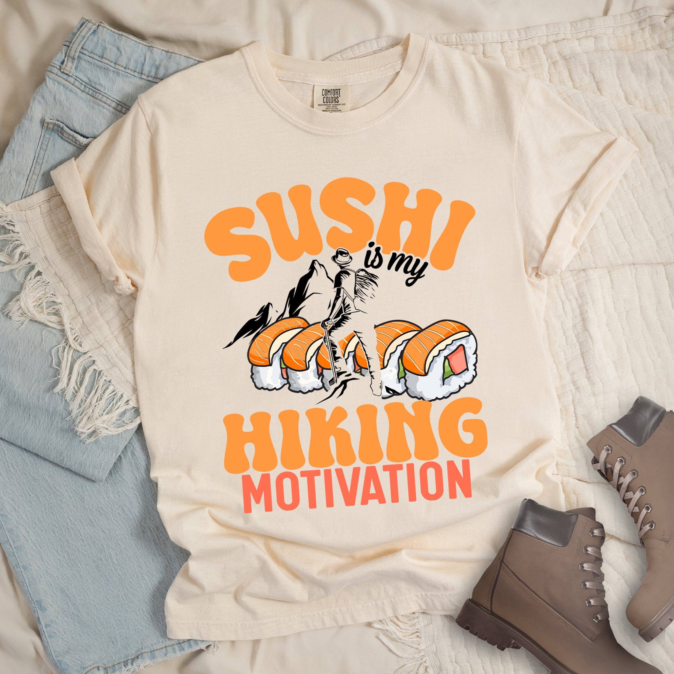 Ivory (Cream White) t-shirt with orange and black text reading "SUSHI is my HIKING MOTIVATION". "SUSHI" appears in large orange letters at the top, "is my" in small black text, and "HIKING" in orange with "MOTIVATION" in red at the bottom. Features a humorous illustration of a hiker climbing over giant sushi rolls, with black mountain silhouettes in the background.