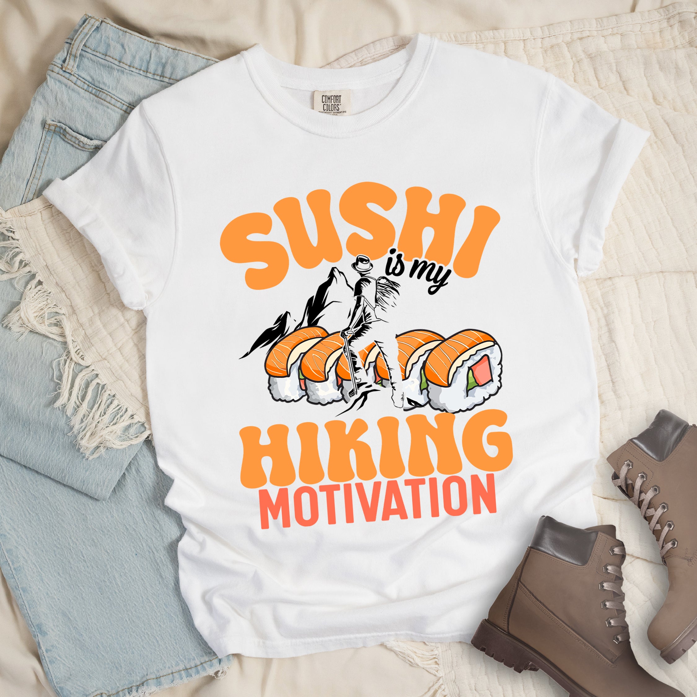 White t-shirt with orange and black text reading "SUSHI is my HIKING MOTIVATION". "SUSHI" appears in large orange letters at the top, "is my" in small black text, and "HIKING" in orange with "MOTIVATION" in red at the bottom. Features a humorous illustration of a hiker climbing over giant sushi rolls, with black mountain silhouettes in the background.