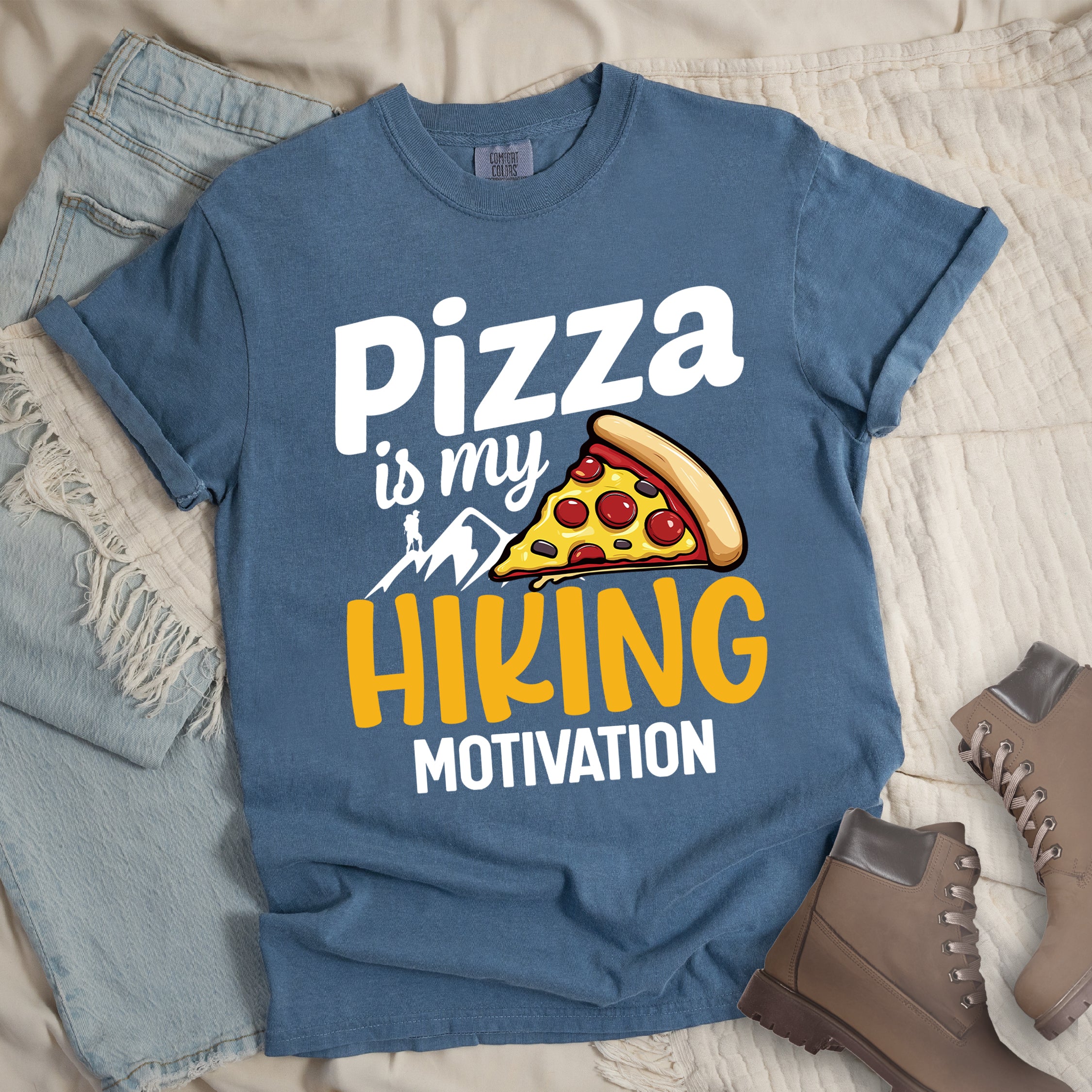 Blue Jean (Denim Blue) t-shirt with white and yellow text reading "Pizza is my HIKING MOTIVATION". "Pizza" appears in large white letters at the top, "is my" in smaller white script, "HIKING" in yellow at the bottom, and "MOTIVATION" in white. Features a colorful pizza slice with pepperoni toppings, white mountain silhouettes with a small hiker figure on a peak. The design humorously connects pizza as the incentive for hiking activities.